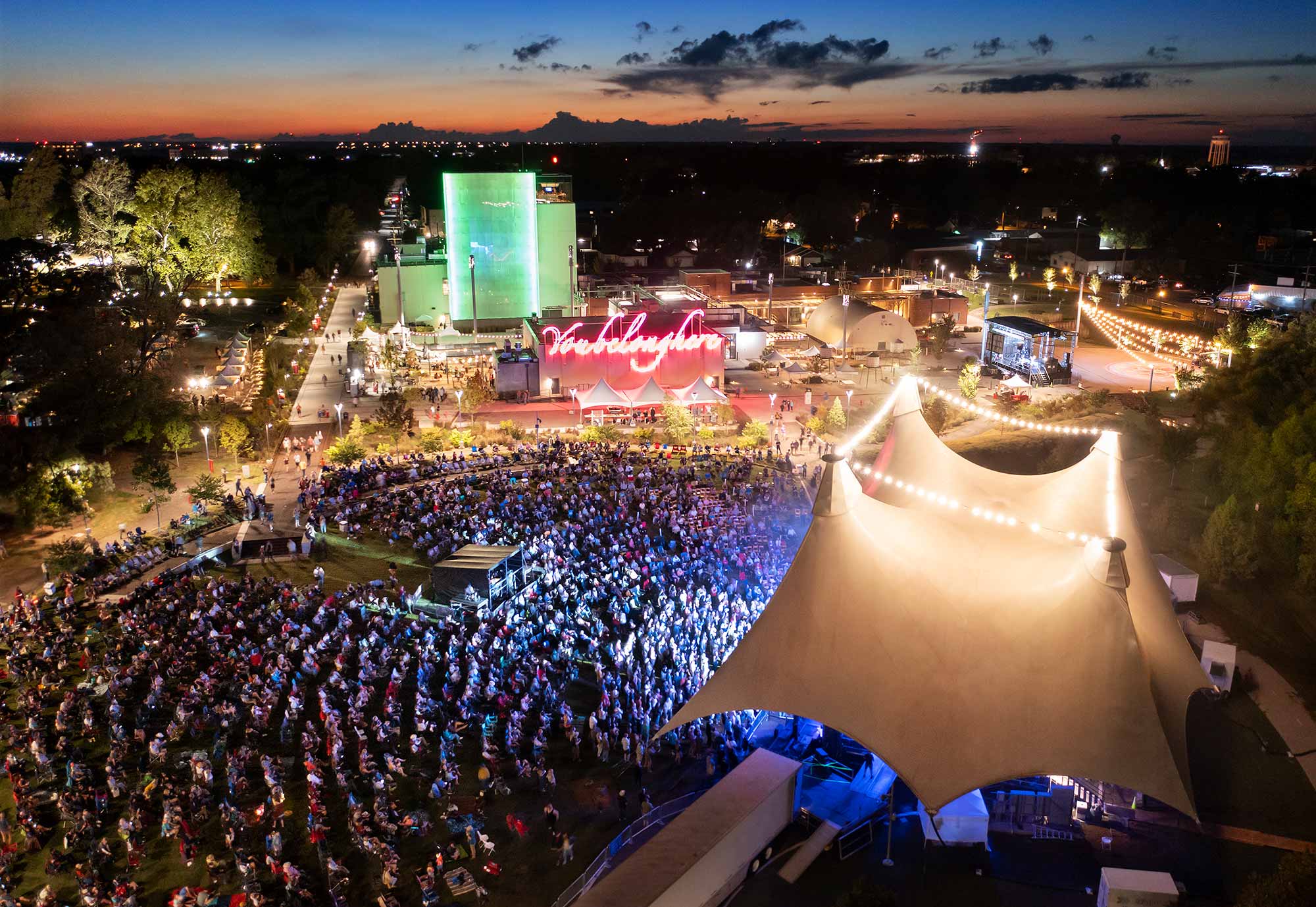 an aerial view of the Momentary at night with the canopy lit with string lights and thousands of people on the green listening to a concert