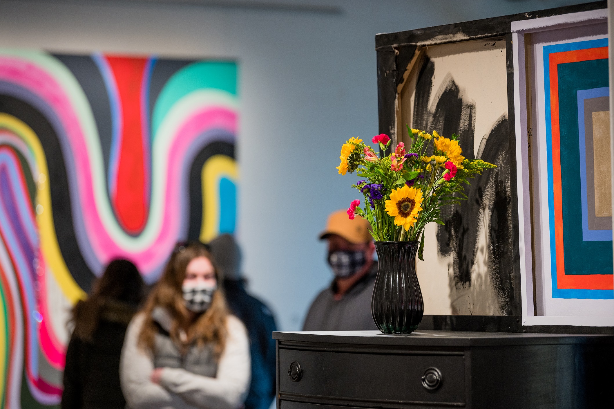 A vase of sunflowers sits on top of a black dresser in the foreground; a large multicolored abstract painting can be seen in the background on a wall