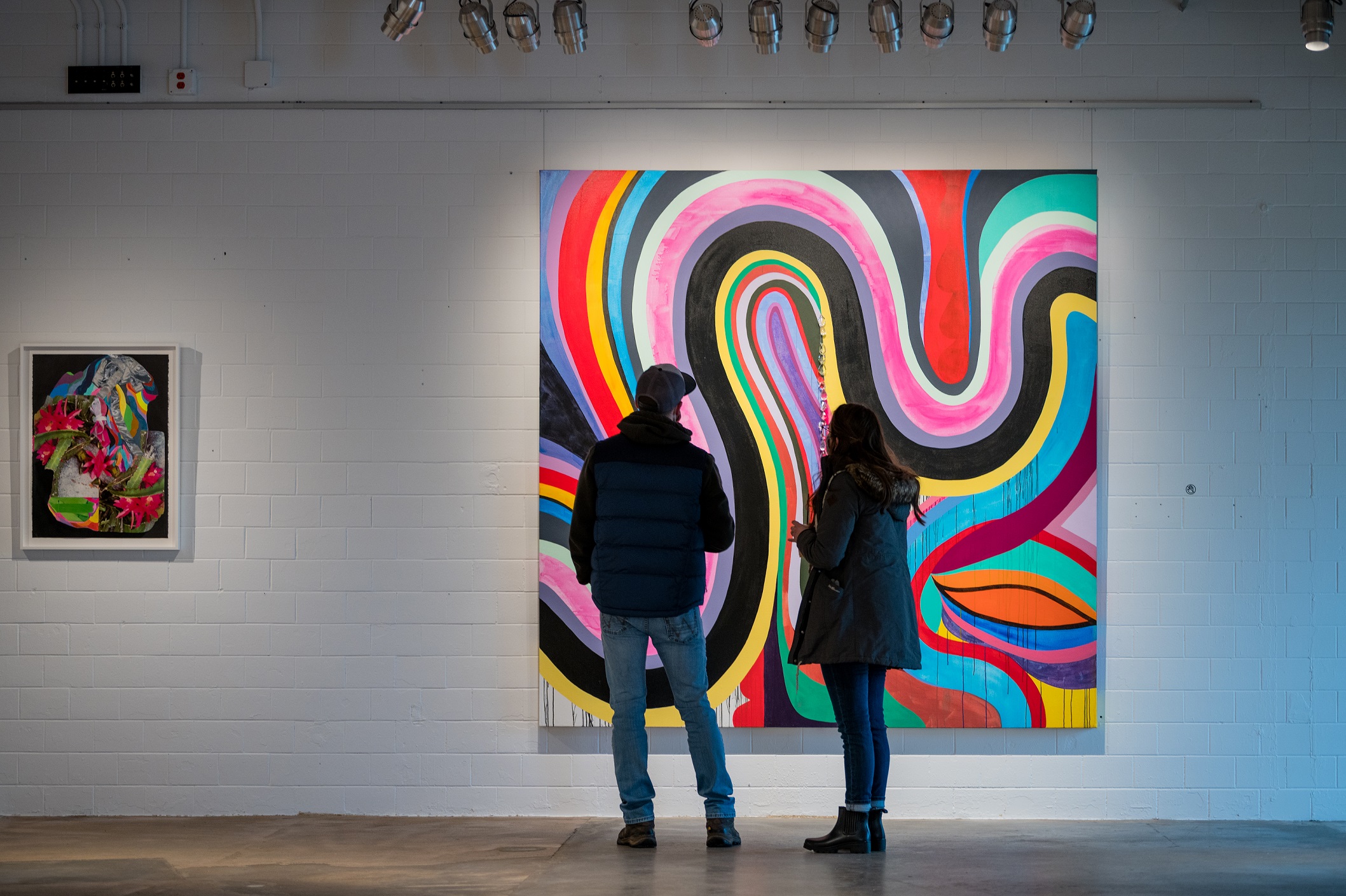 Two people looking at a multicolored abstract painting with large loops and swirls on the wall