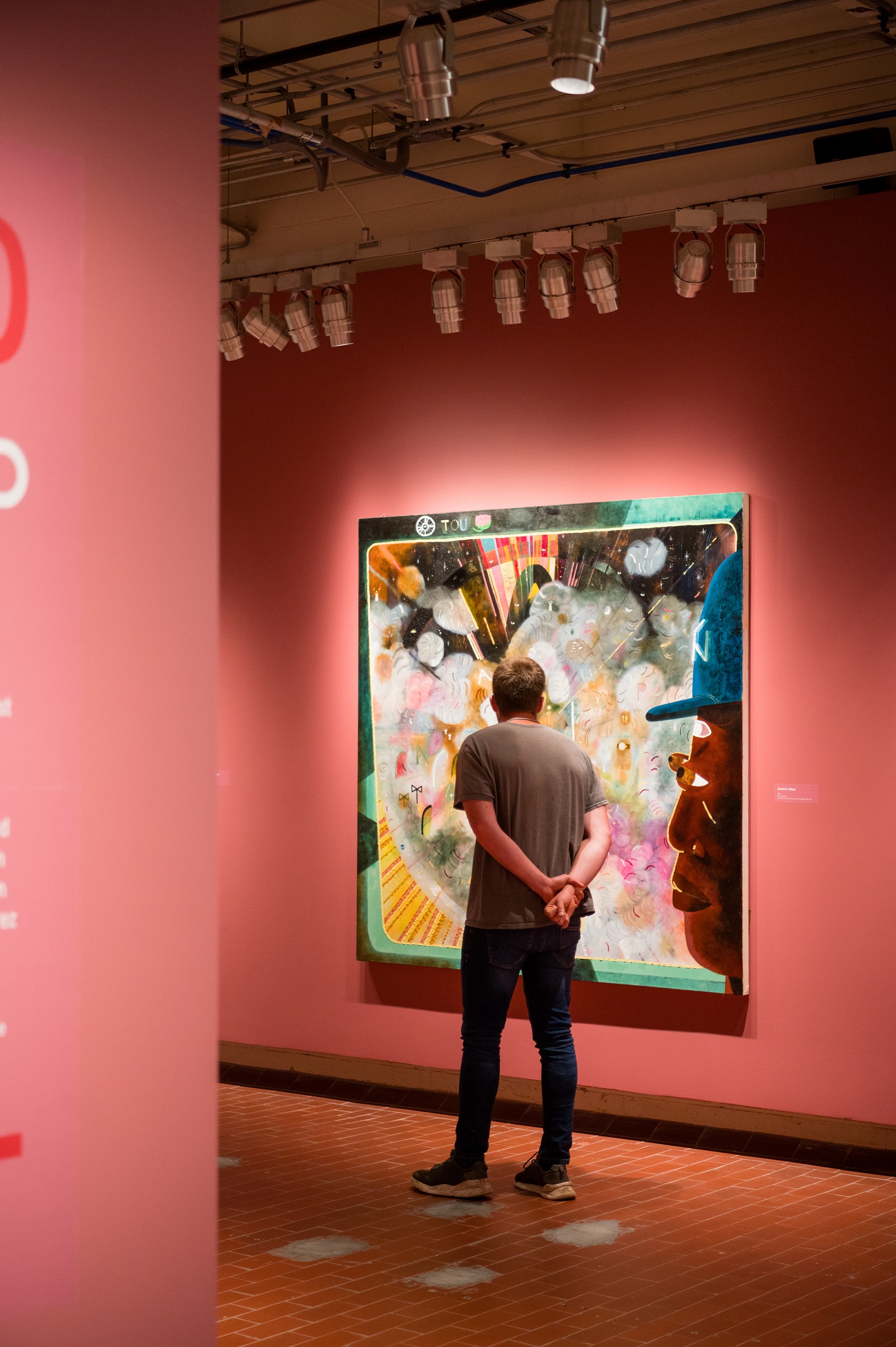 A man stares at a large painting on a wall with many colors and the face of a black man looking on from the right side of the painting