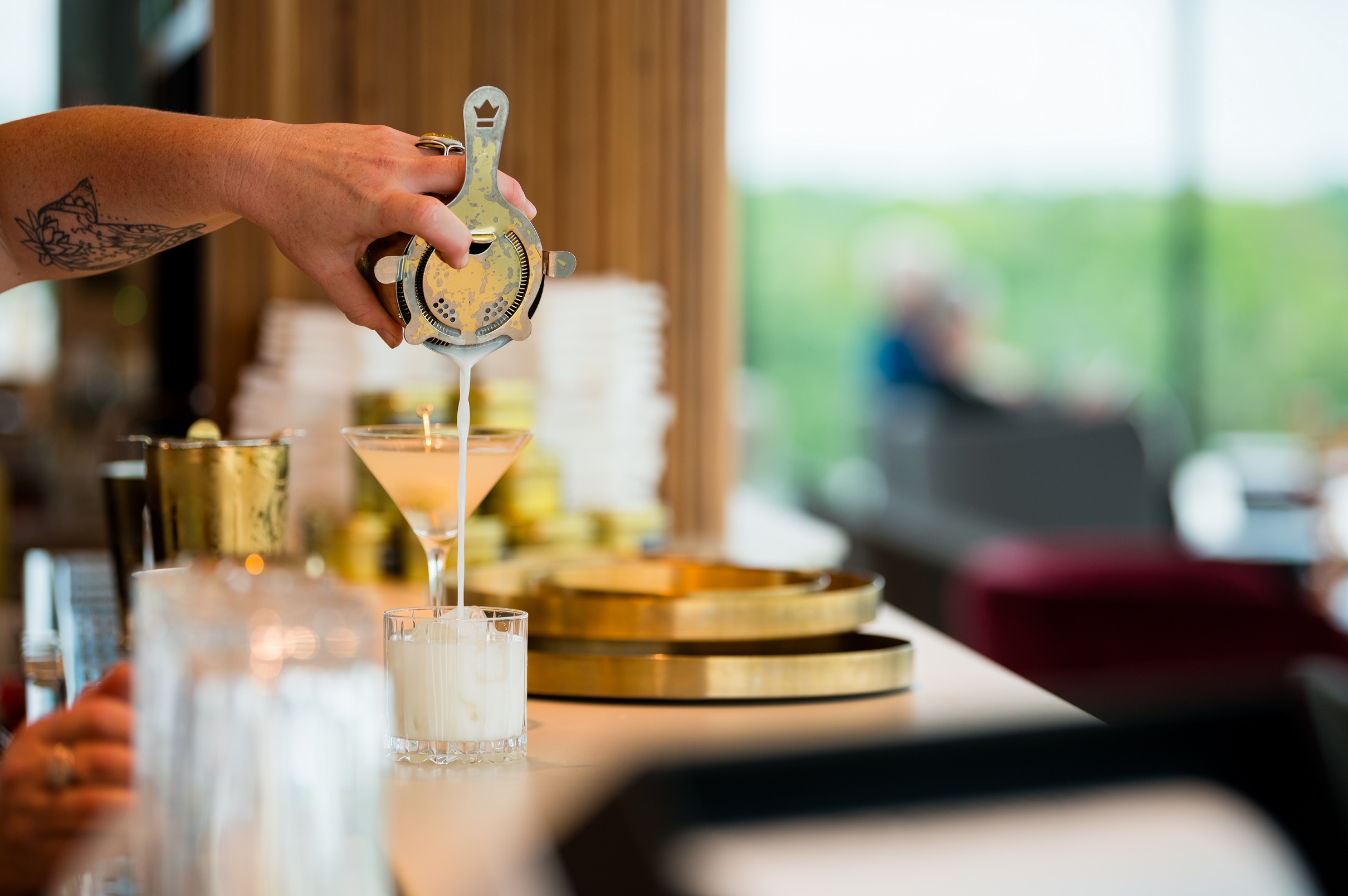 A bartender pours a drink from a gold shaker into a glass on the bar