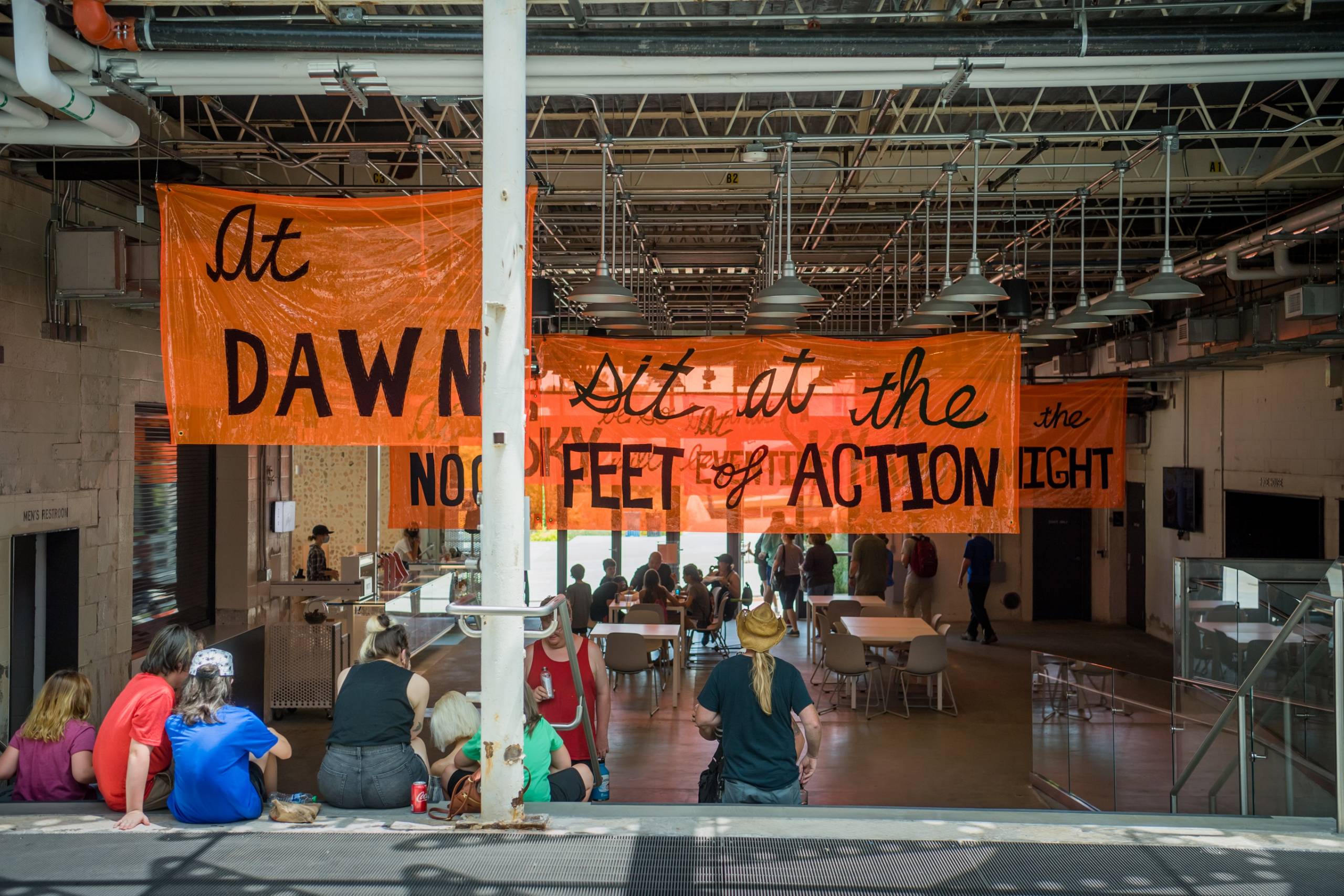People sitting on steps inside with large orange banners overhead with words and phrases written in black