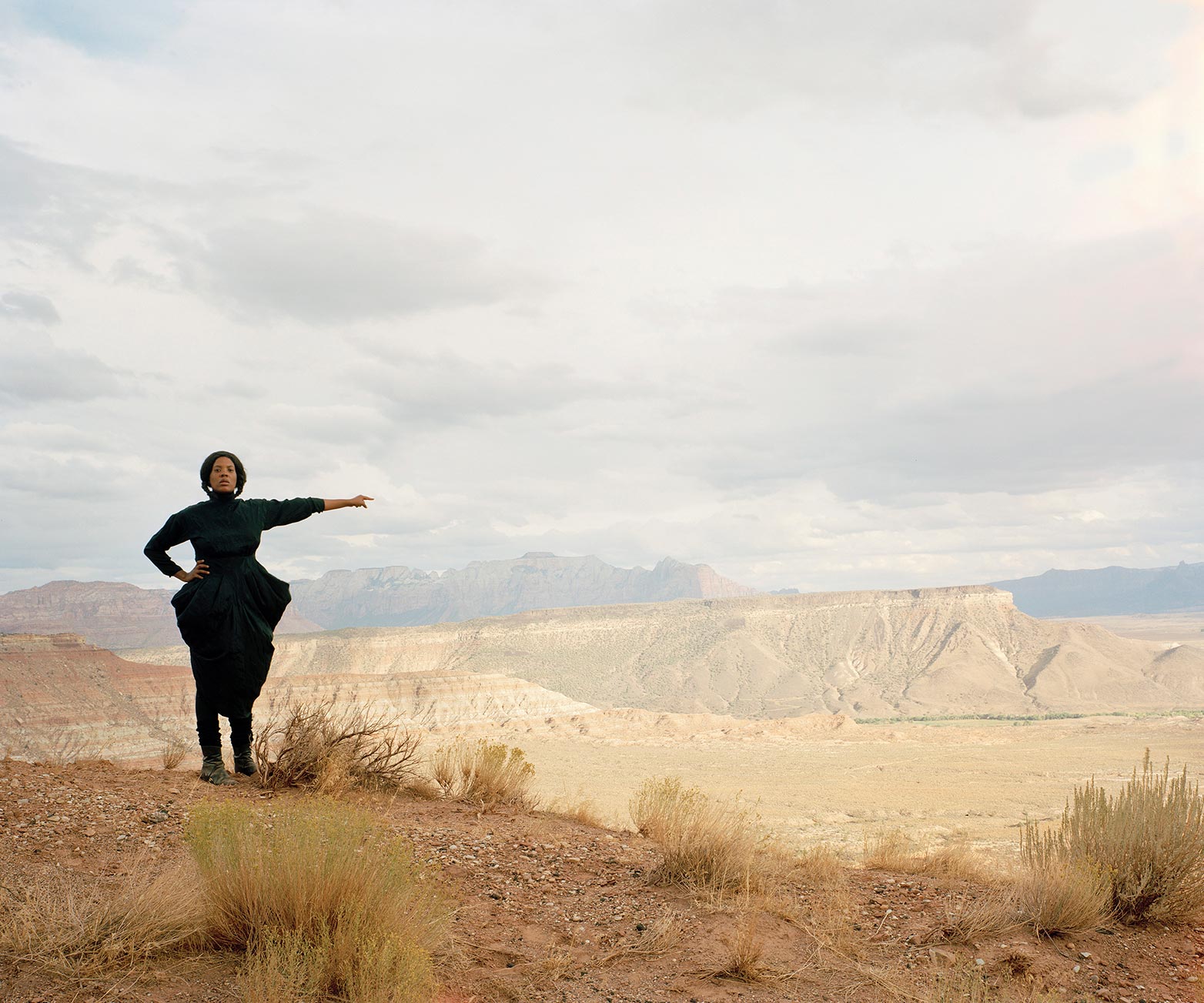A person in a black dress stands on a desert vista pointing to the landscape against clouds and dusty mountains