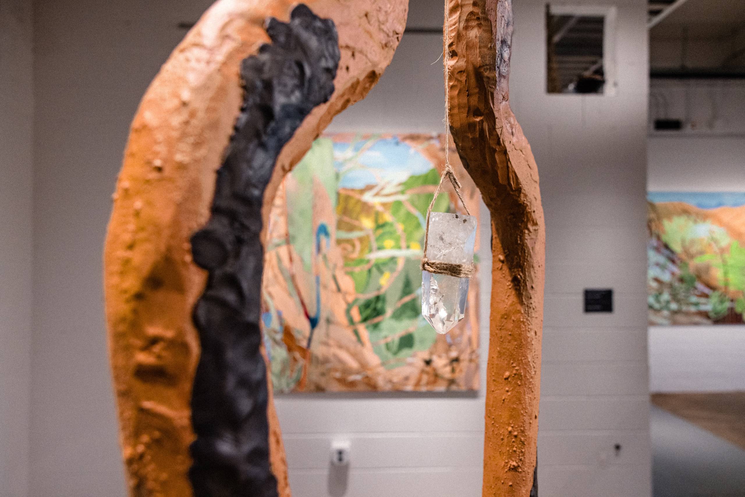 A crystal hangs from a rope inside an orange and black sculpture that frames an abstract painting in the background with browns, oranges, greens, blues, and yellows.
