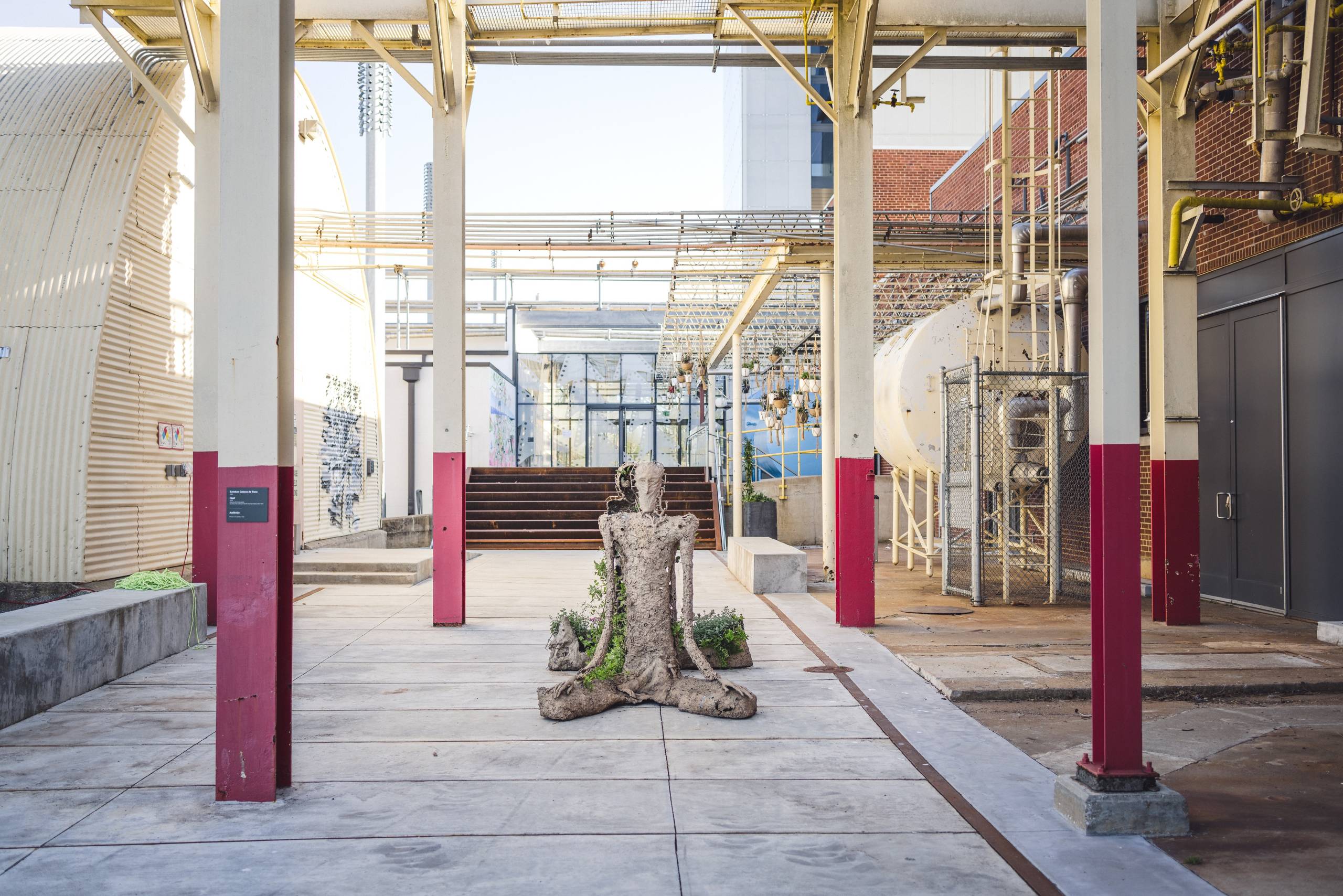 A sculpture of a cross-legged figure with plants and greenery around it sitting between red and tan beams and a boiler outside of the Momentary.