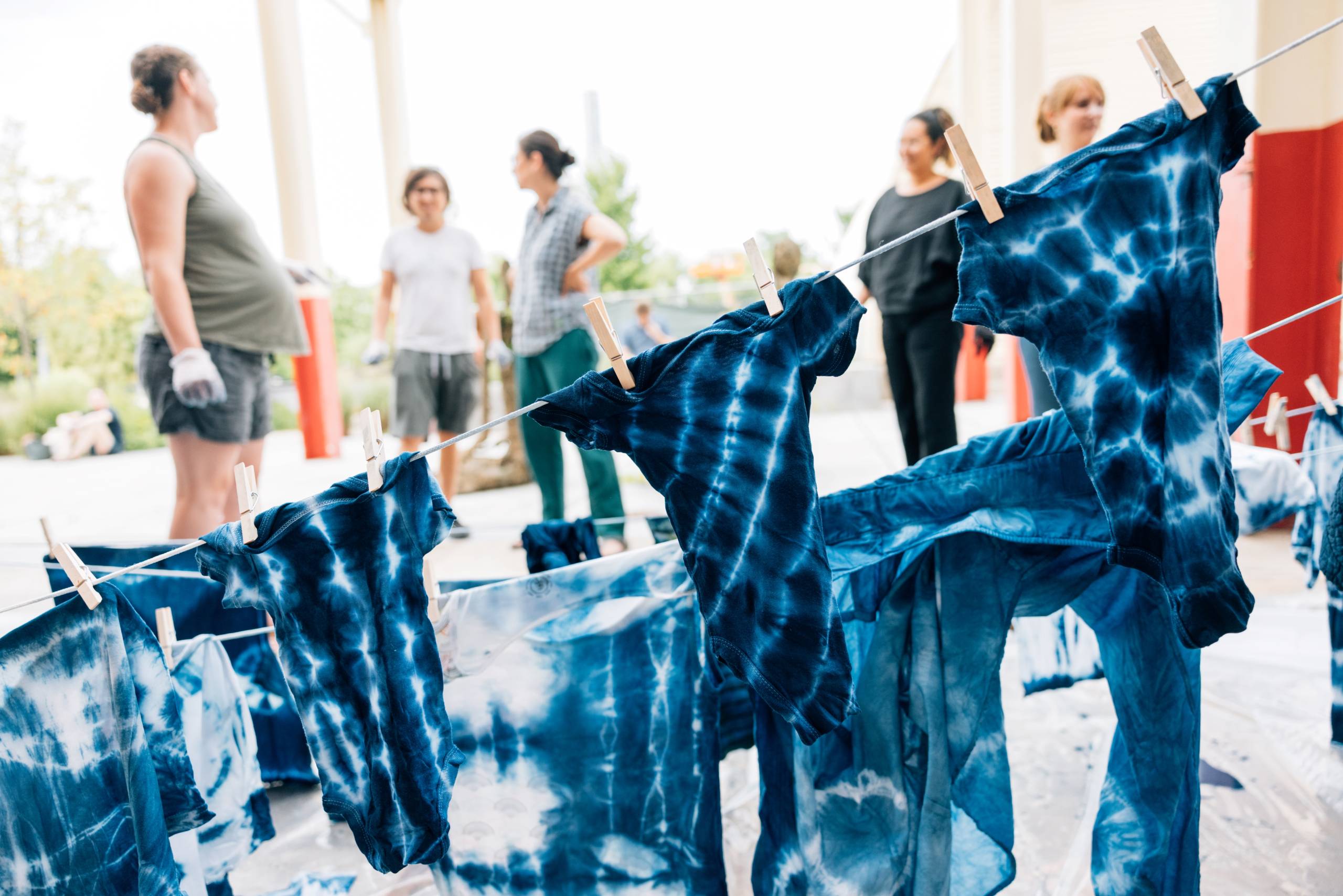 Freshly dyed clothing dries on a clothesline, while several people talk happily in the background