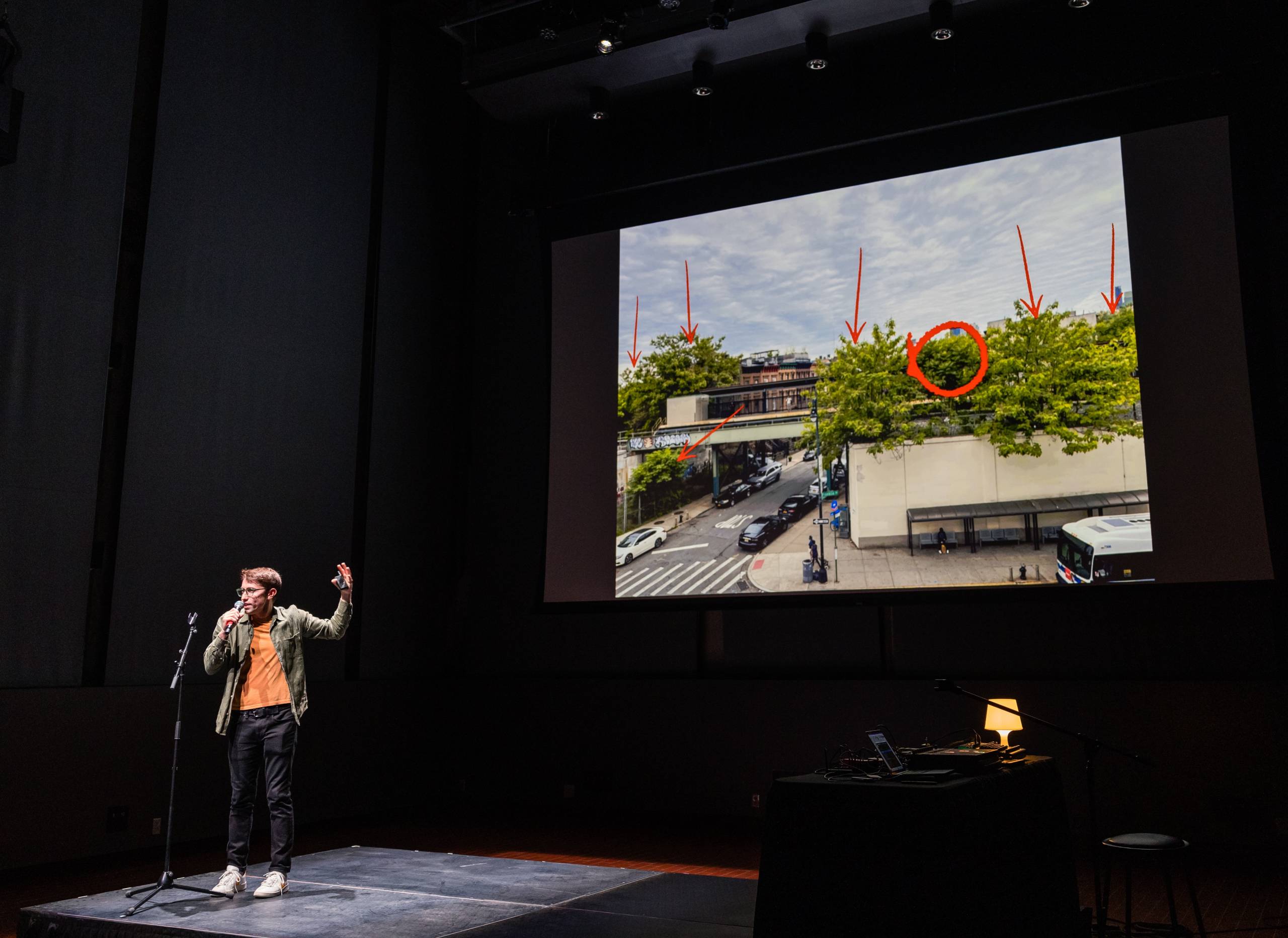 a young white man in glasses holds a microphone on stage and points behind him to a screen featuring an urban scene and arrows pointing to paulownia trees growing through cement