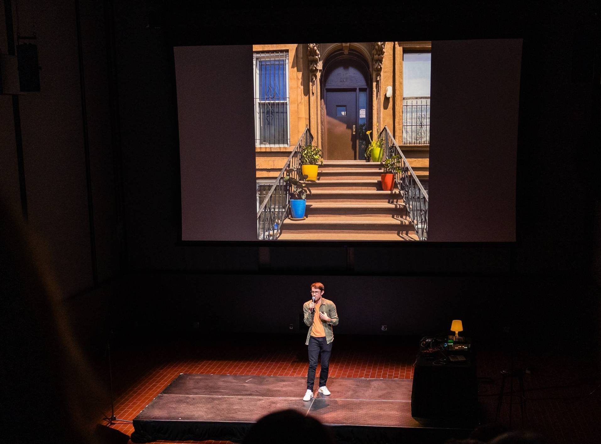 a young white man in glasses holds a microphone on stage while a projector behind him shows an image of a stoop with four brightly colored flower pots, one blue, one red, one green, and one yellow