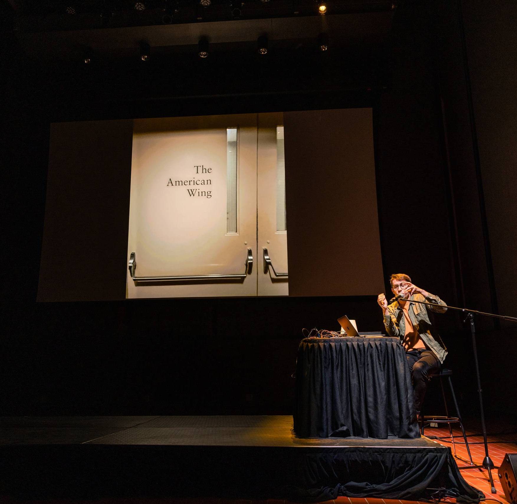 a young white man in glasses speaks into a microphone on stage while a projector behind him shows an image of a door that says 