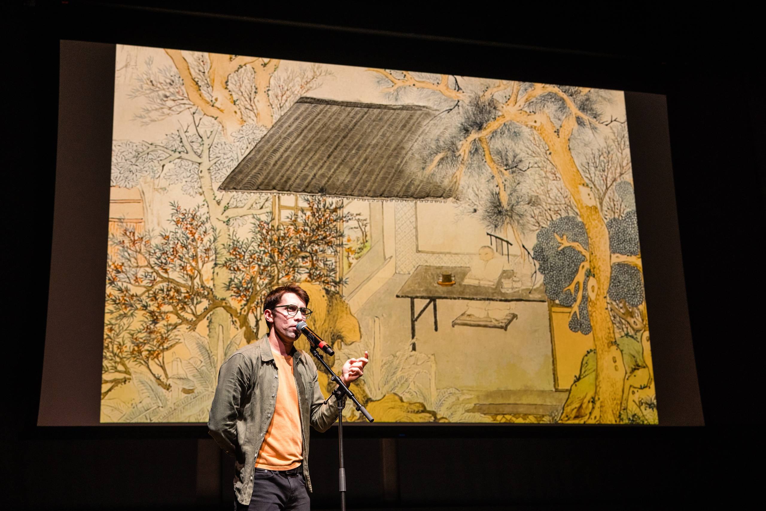 a young white man in glasses speaks into a microphone while a projector behind him shows an image of a sixteenth-century Chinese painting of a man playing a synth for his pet crane under a paulownia tree