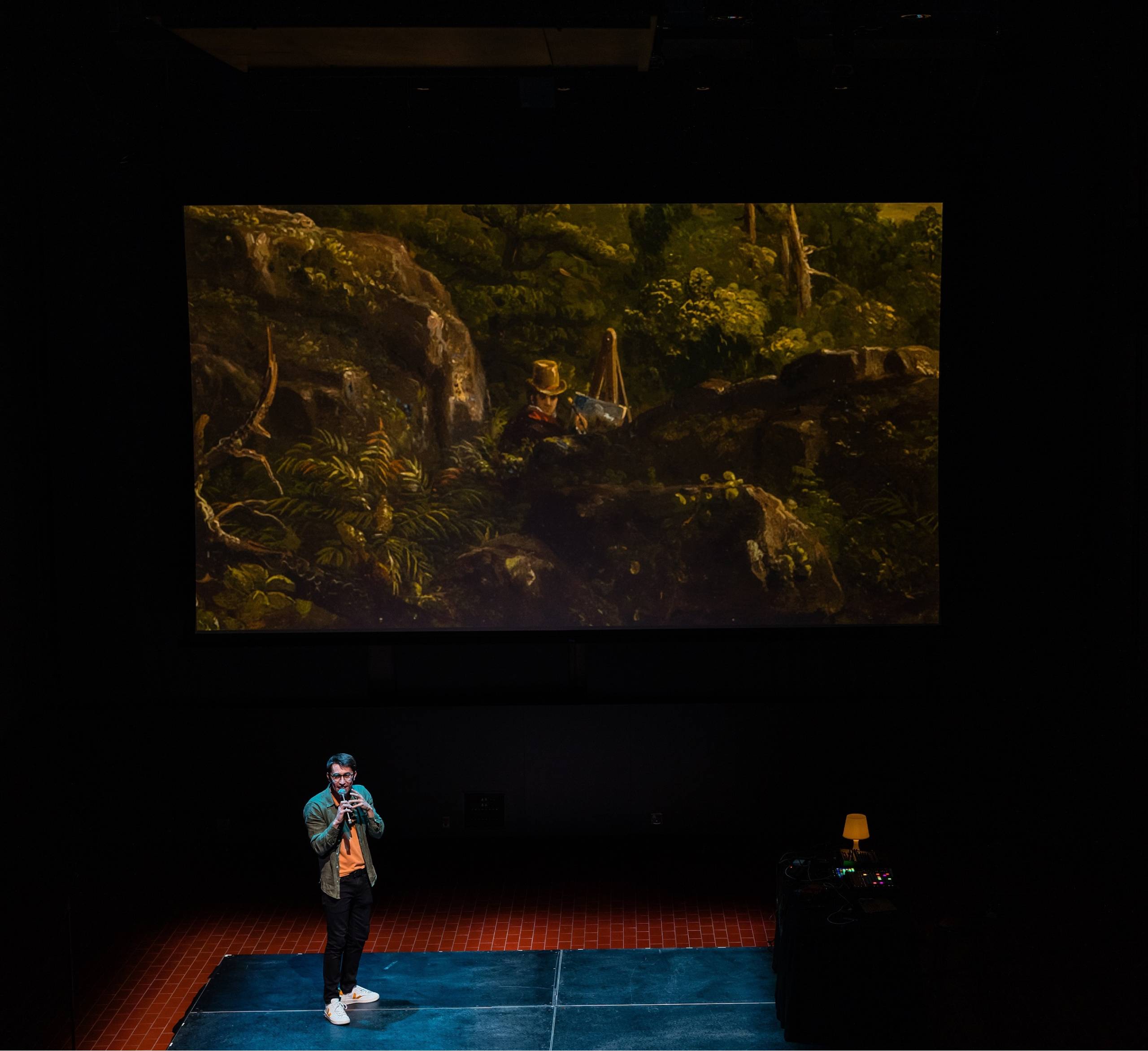 a young white man in glasses holds a microphone on stage while a projector shows detail of a Thomas Cole painting where the artist has painted himself into the scene