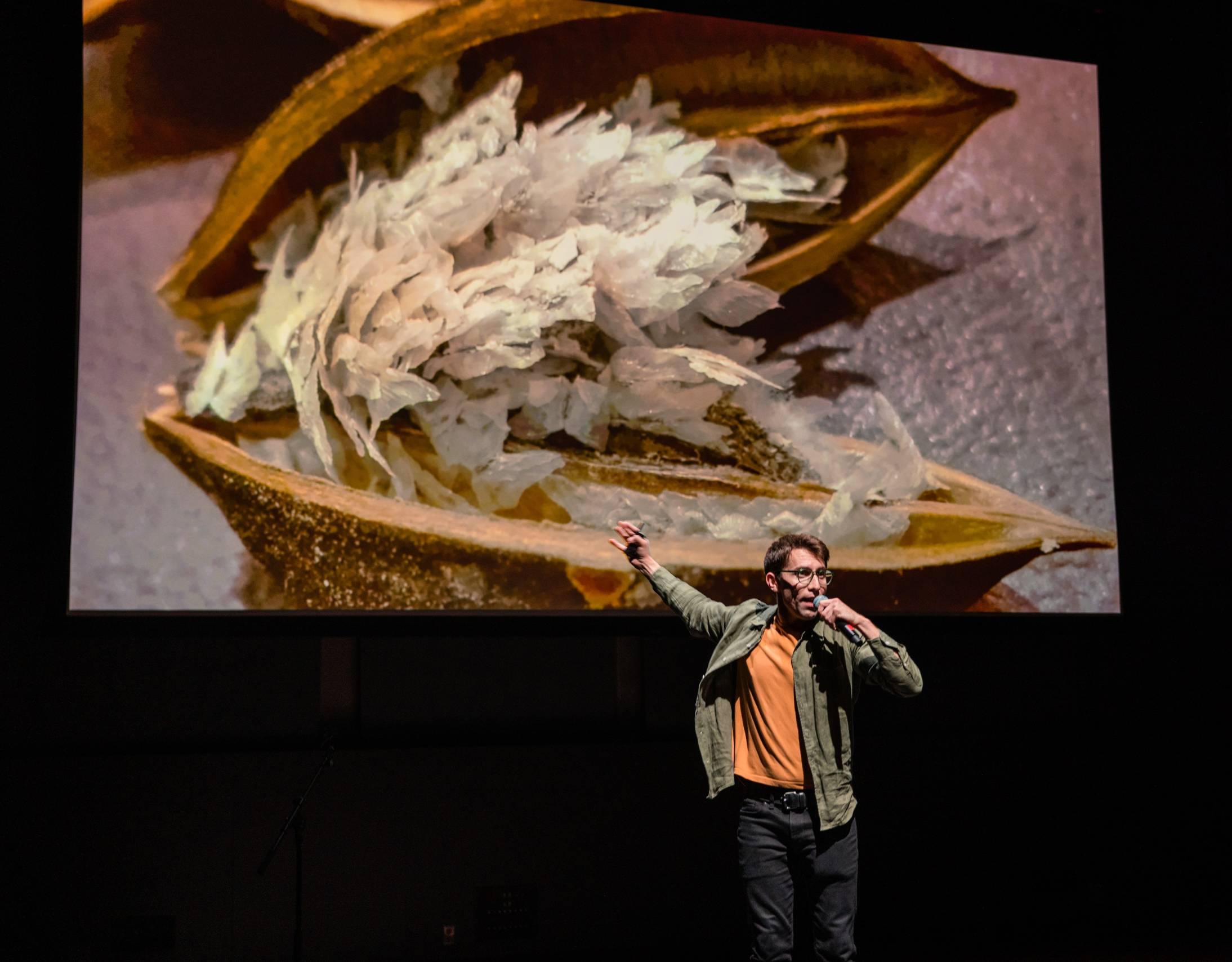 a young white man in glasses holds a microphone on stage and points behind him to a screen featuring an open paulownia seed