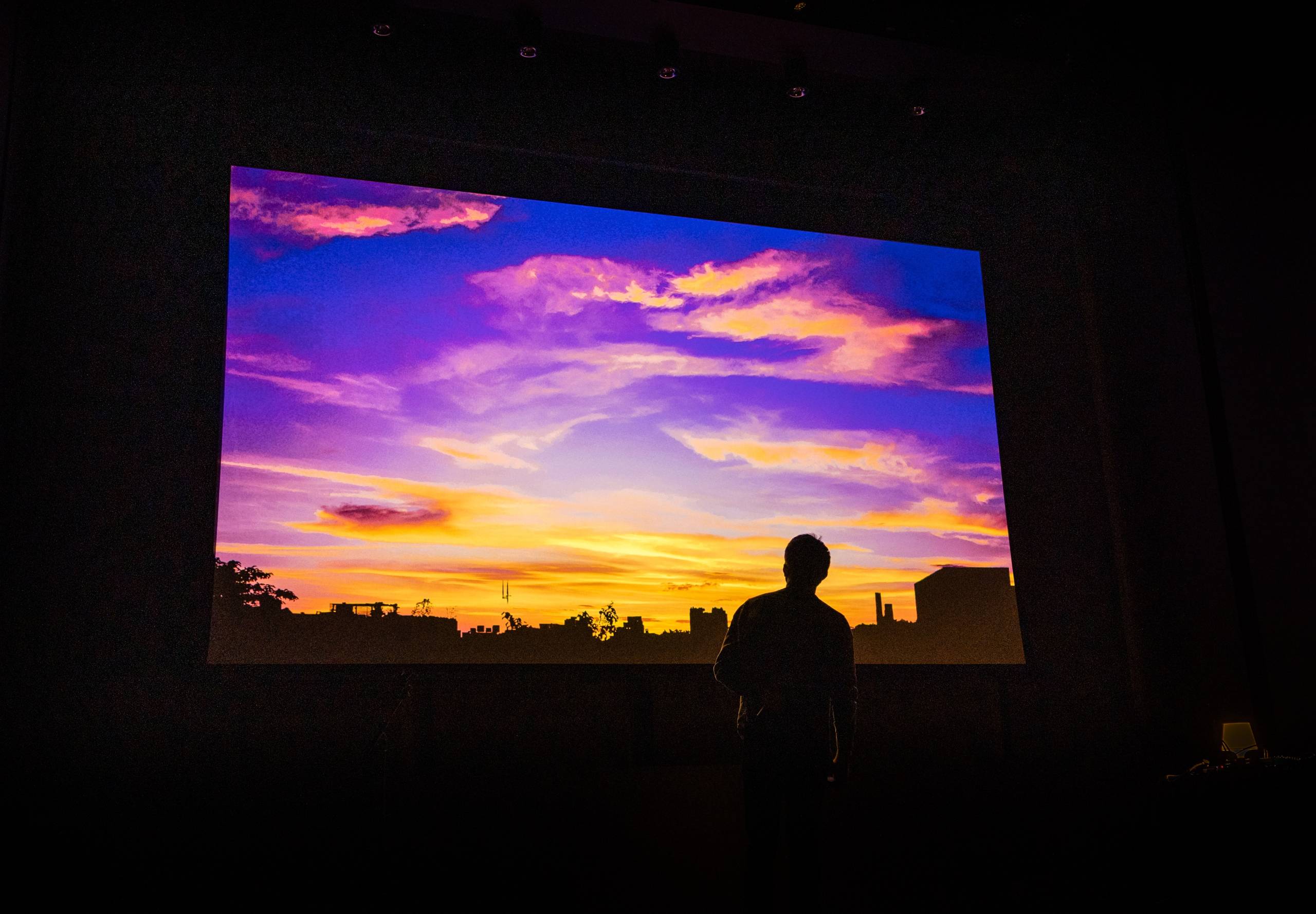 a projected screen shows an image of a sunset in Brooklyn with vibrant shades of blue, purple, orange, and yellow foregrounded by the silhouette of a person staring at it