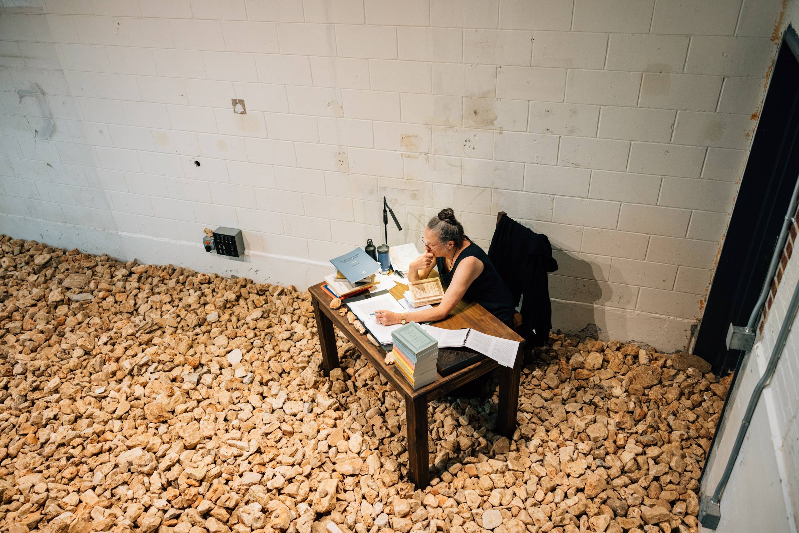 a woman wearing glasses sits at a desk crowded with books reading in a room in which the floor is covered with rocks