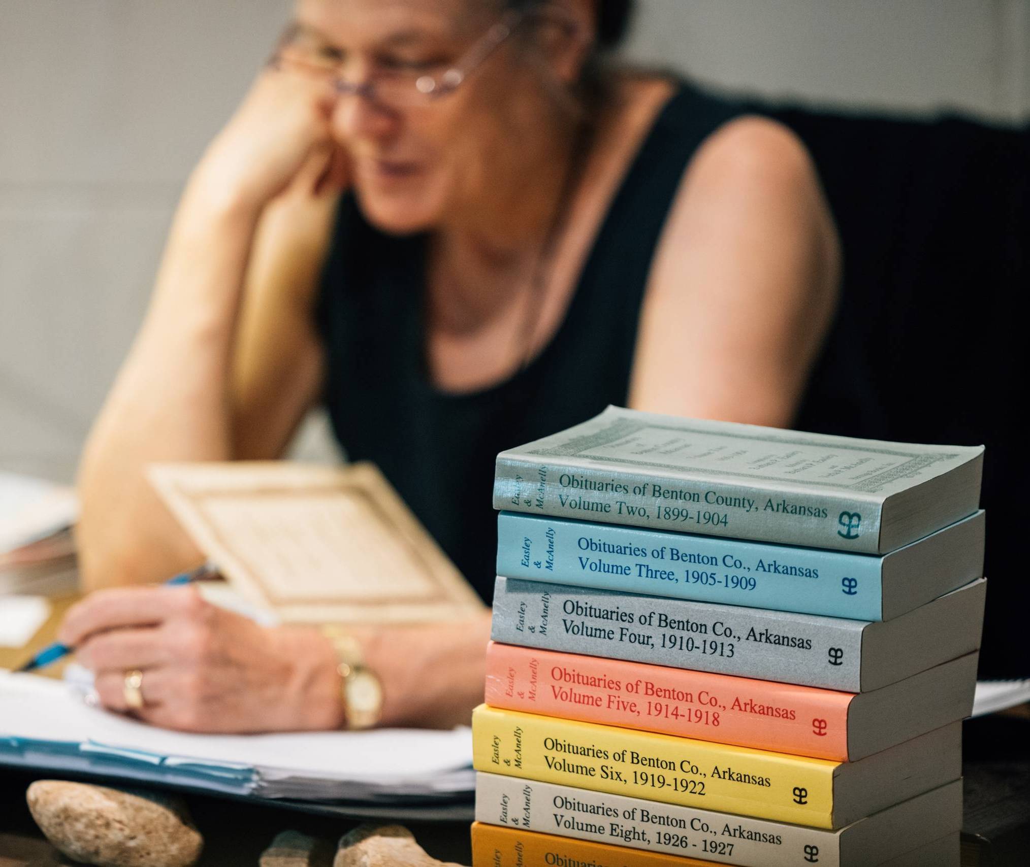 a stack of colorful volumes of obituaries of Benton County foregrounds a woman wearing glasses reading in the background