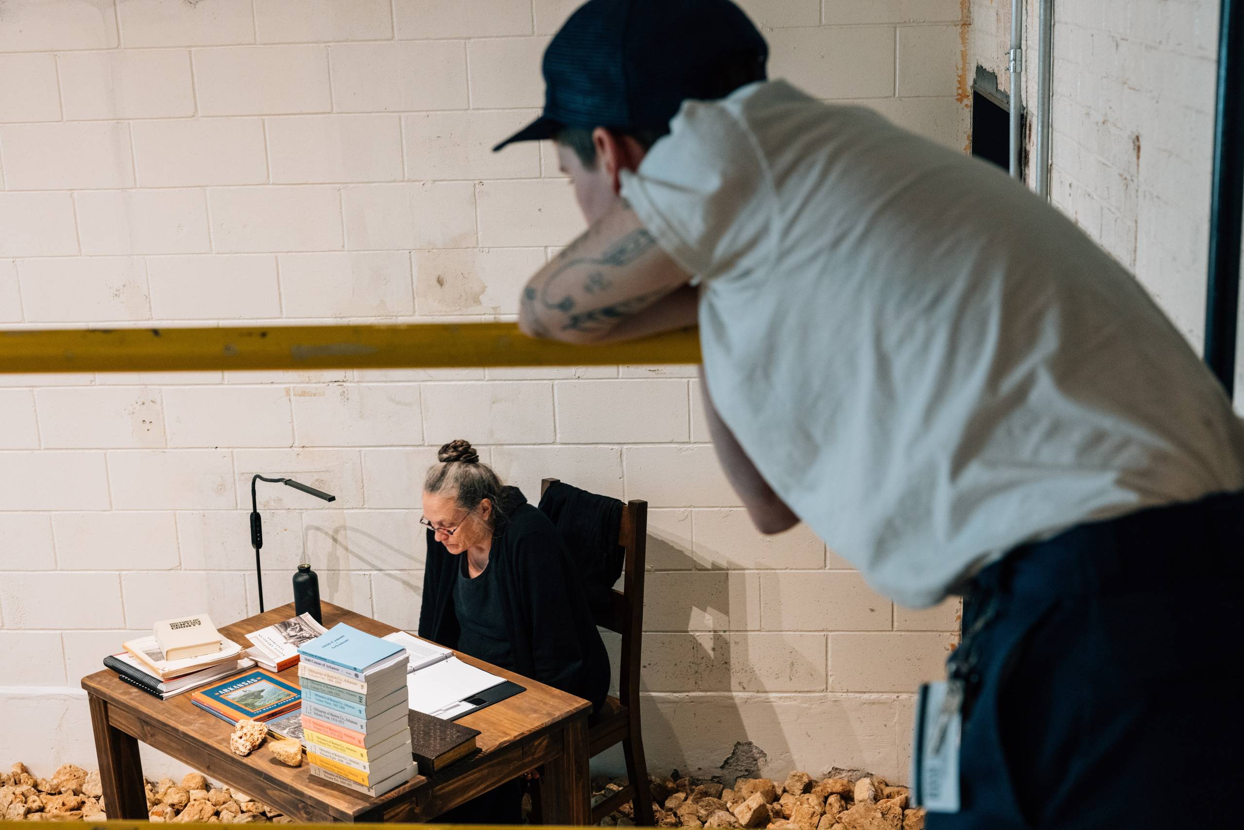 a young individual rests their chin on a balcony rail watching a woman wearing glasses sitting at a desk and reading