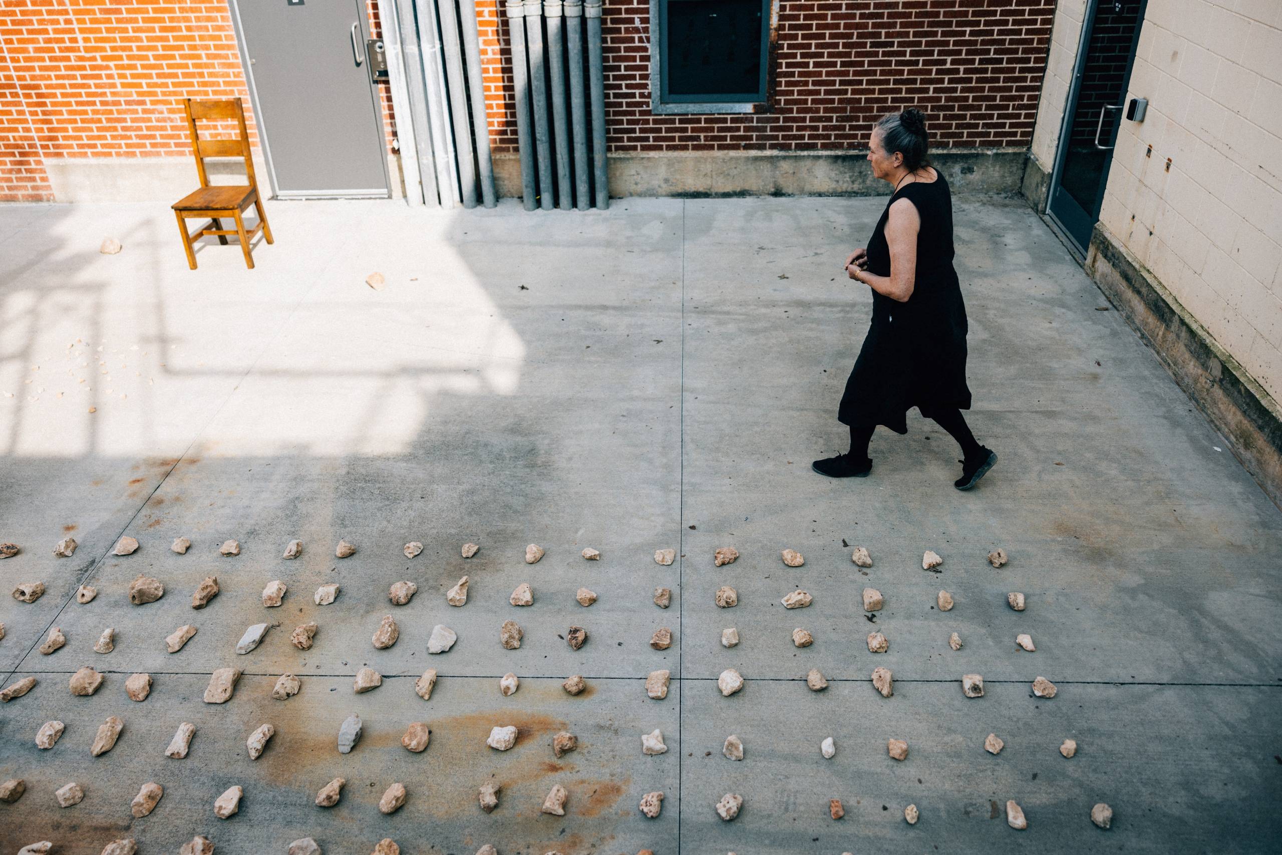 a woman wearing black walks across a cement floor organizing rocks into rows