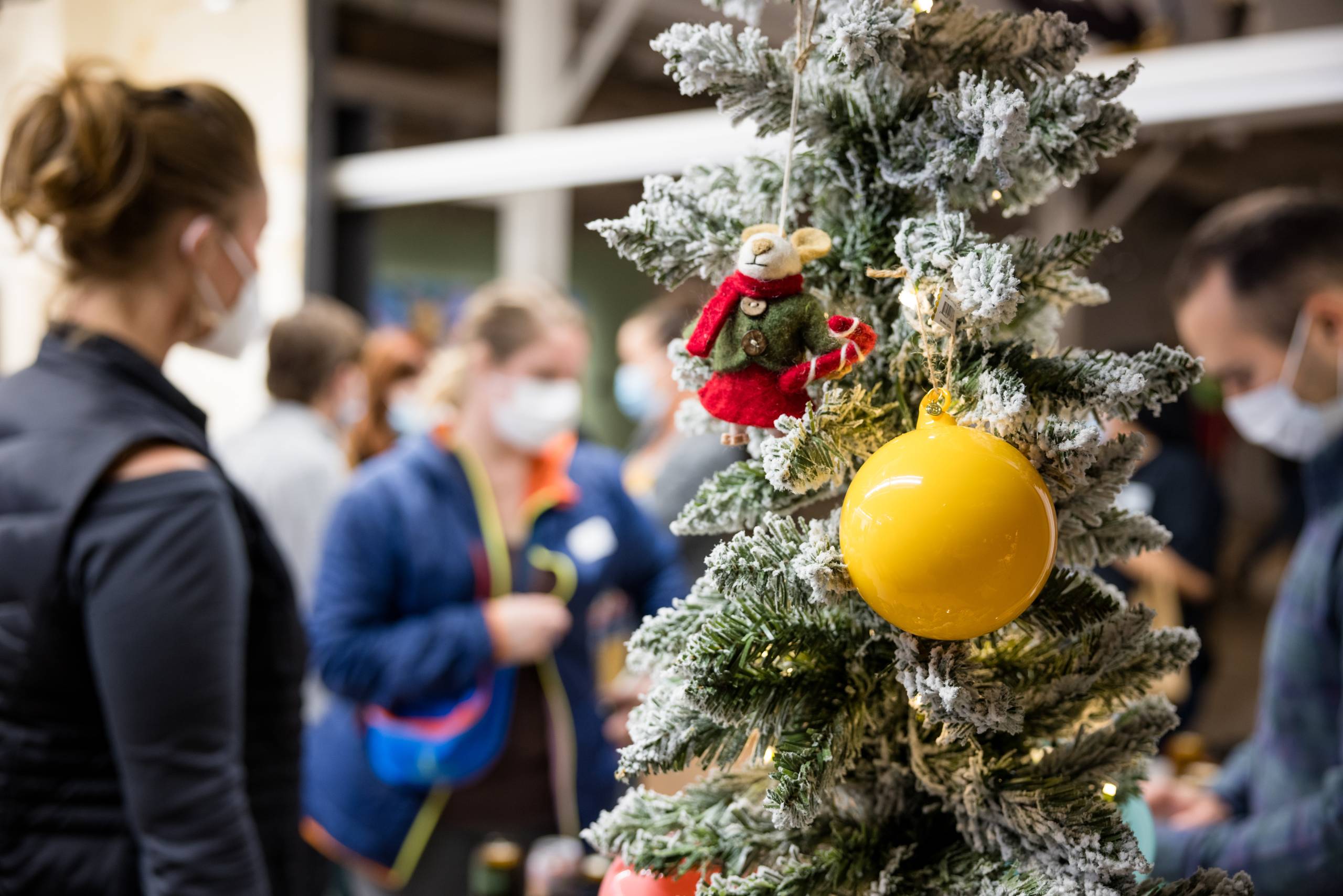a christmas tree decorated with a yellow ball and a mouse ornament foregrounds people mingling in the background