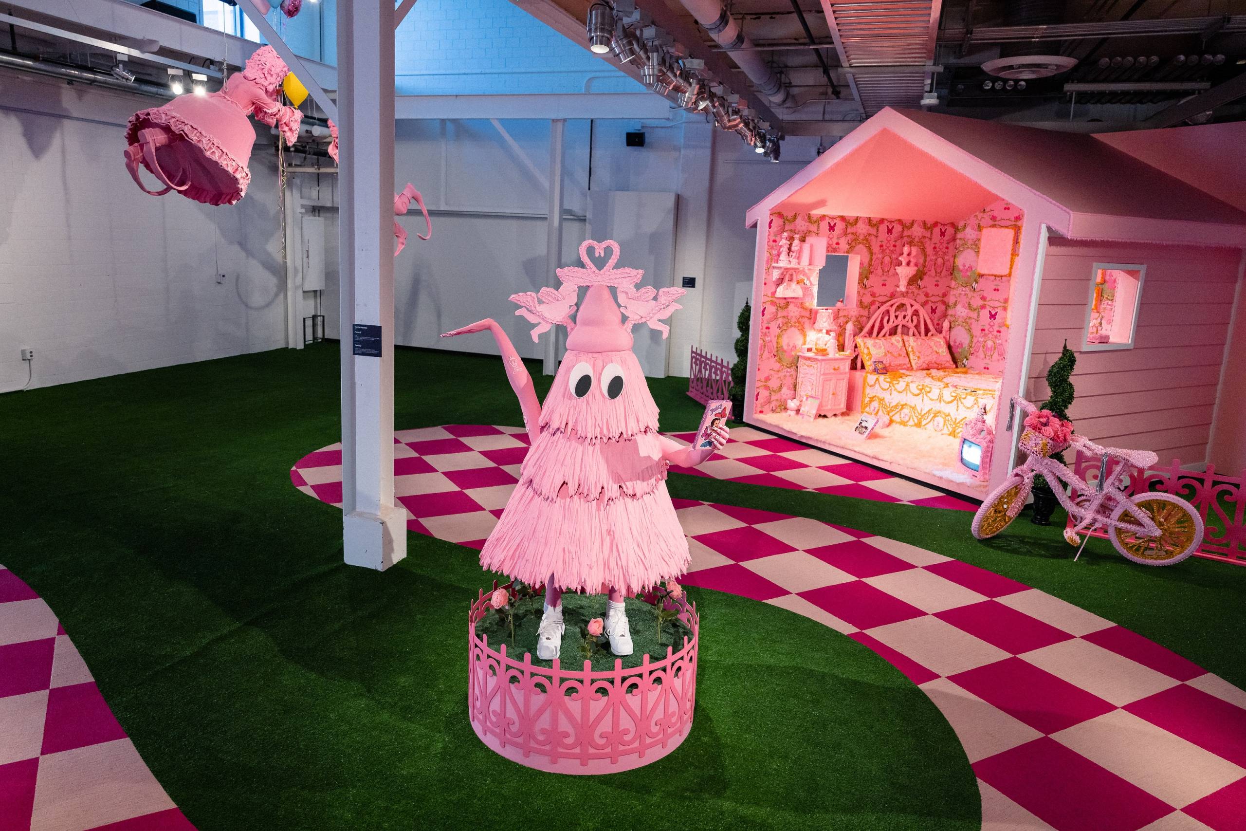 a gallery space with multiple installations in bright shades of pink such as a bedroom, a bicycle, a treelike figure holding a phone, and a checkered path through a green lawn floor