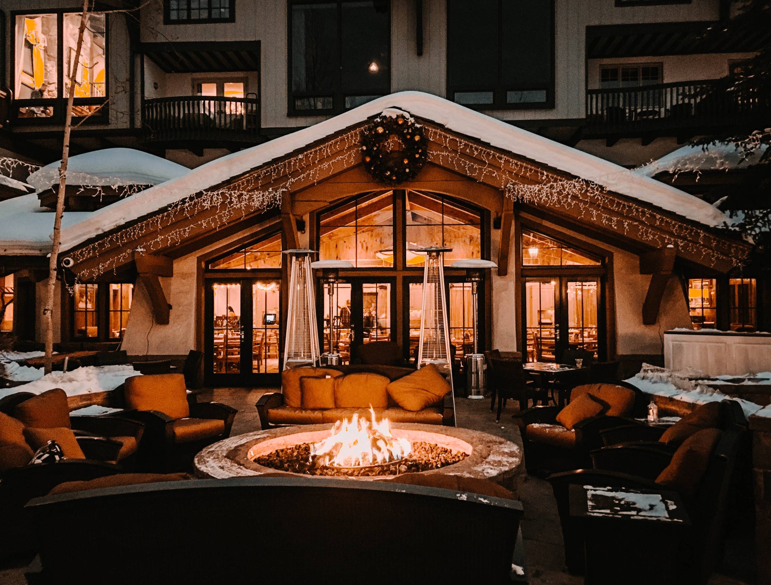 A ski lodge with a small amount of snow on its roof, with a burning fire pit, chairs, and cushions in the foreground