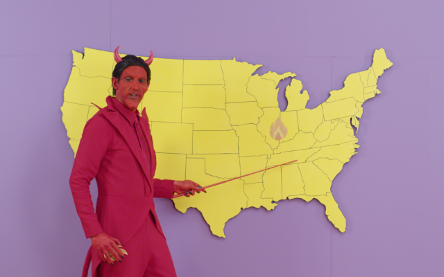 A man in a red devil costume points an indicator wand at a cutout map of the continental United States.