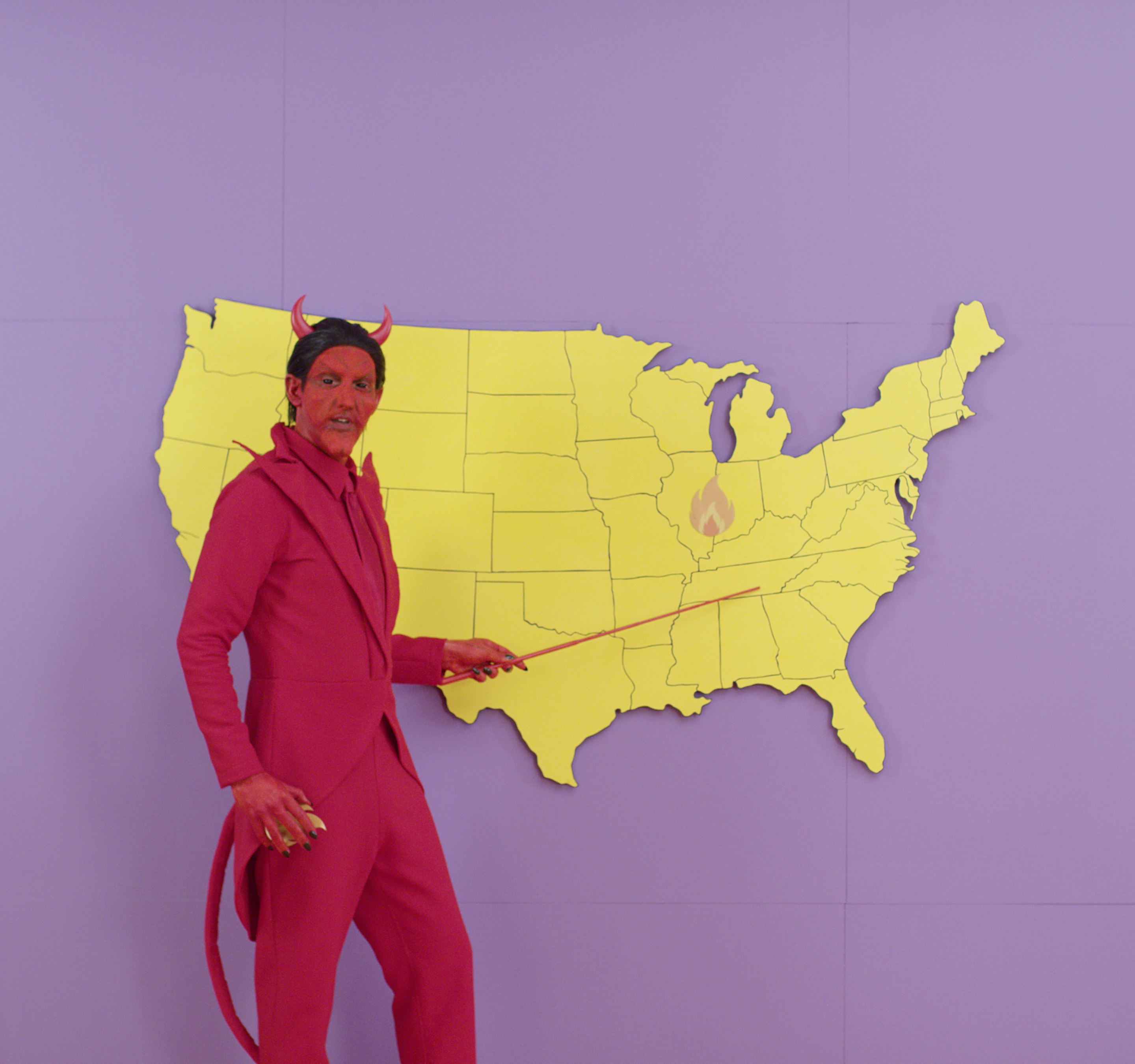 A man in a red devil costume points an indicator wand at a cutout map of the continental United States.