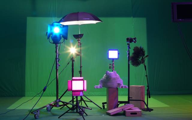 A collection of stage lights, lamps, wooden blocks, and a palm tree stand in front of a fabric green screen on the set of [siccer].