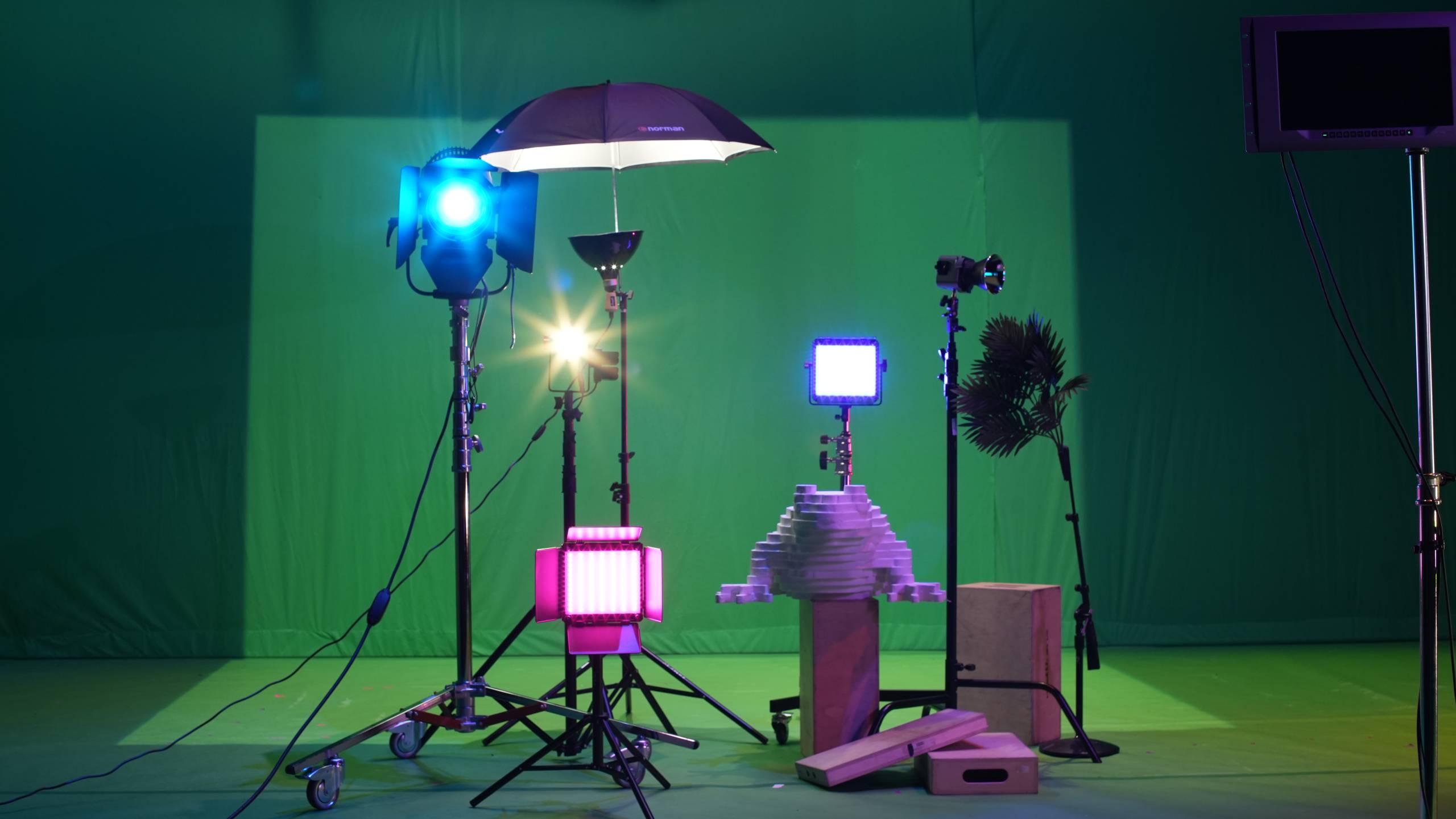 A collection of stage lights, lamps, wooden blocks, and a palm tree stand in front of a fabric green screen on the set of [siccer].