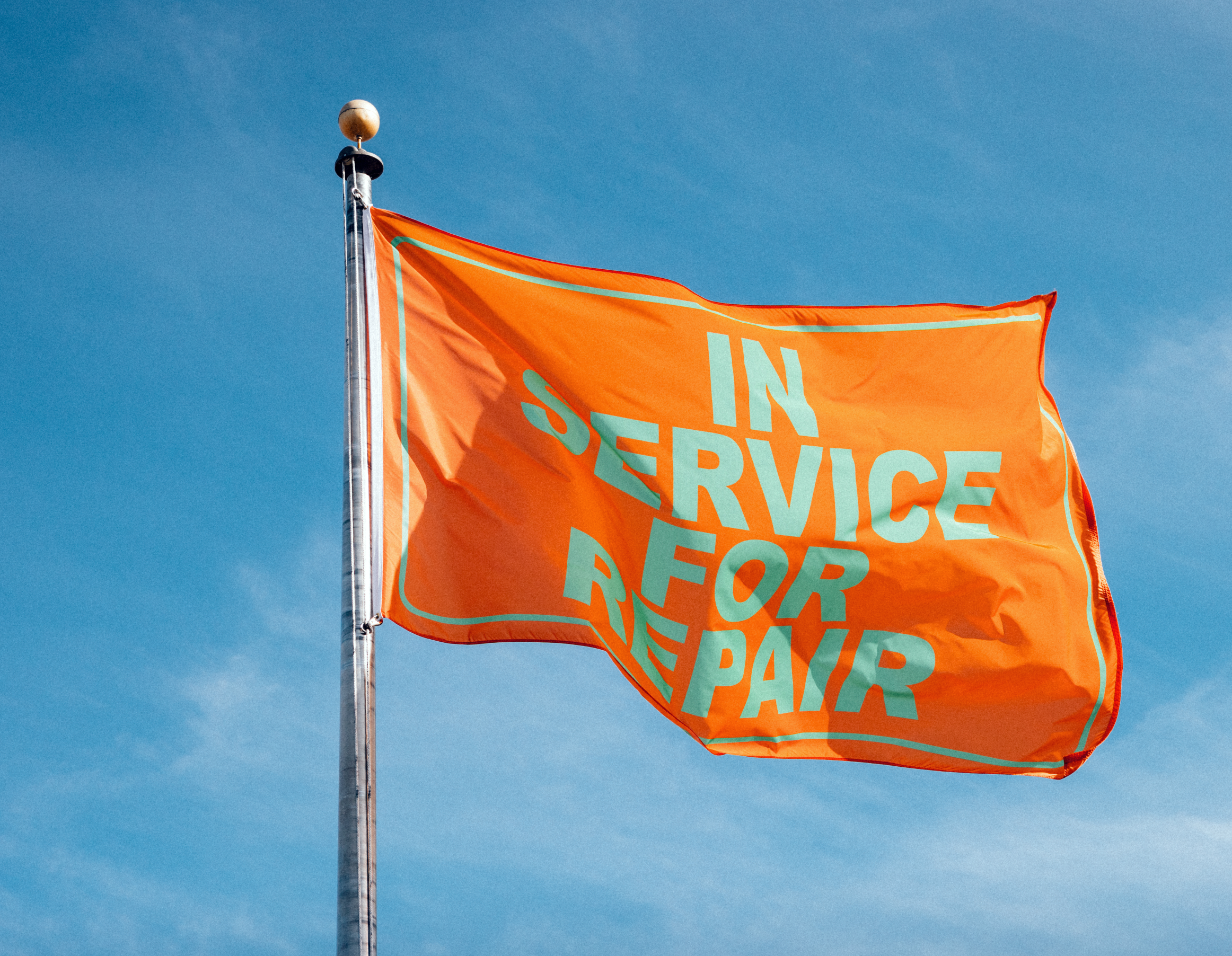 An orange flag waving in the wind, with the words IN SERVICE FOR REPAIR printed in large block letters on the flag.