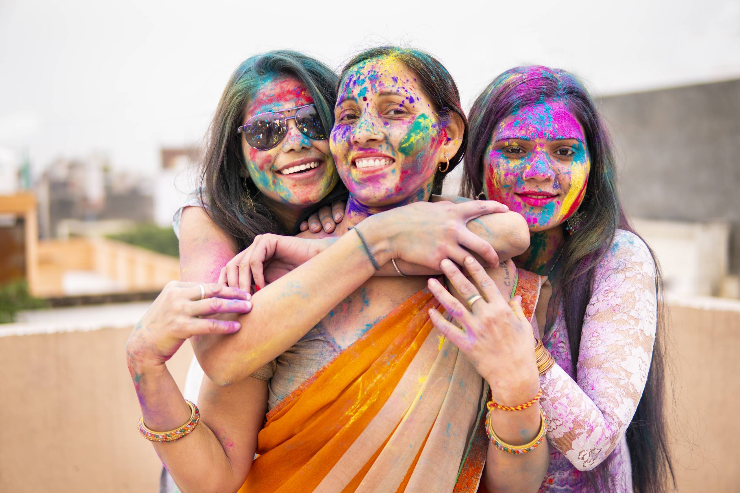 Outdoor image of Asian / Indian happy, beautiful mother daughters in Indian dress celebrating the Holi festival together with color powder. They are standing together and looking at the camera with a toothy smile.