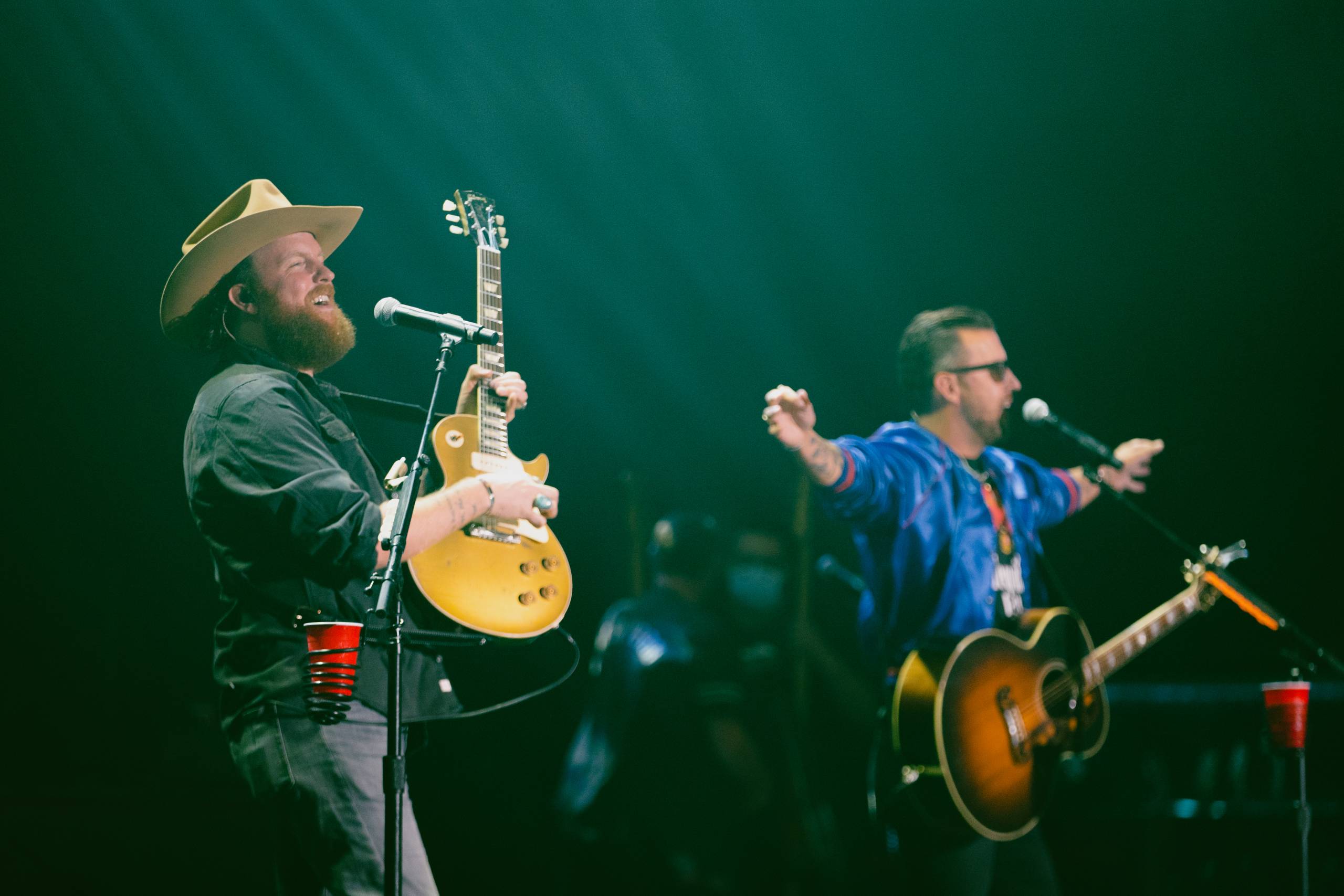 Country duo Brothers Osborne standing and performing on a stage. Both are holding acoustic guitars and singing into standing microphones.
