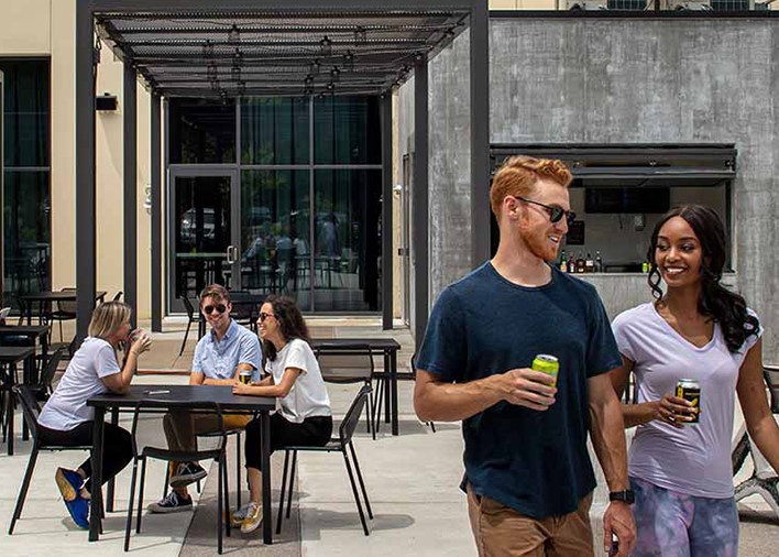 Two people walk across the Arvest bank Courtyard holding drinks and talking, while three young adults sit at a table in the background laughing and holding canned drinks.