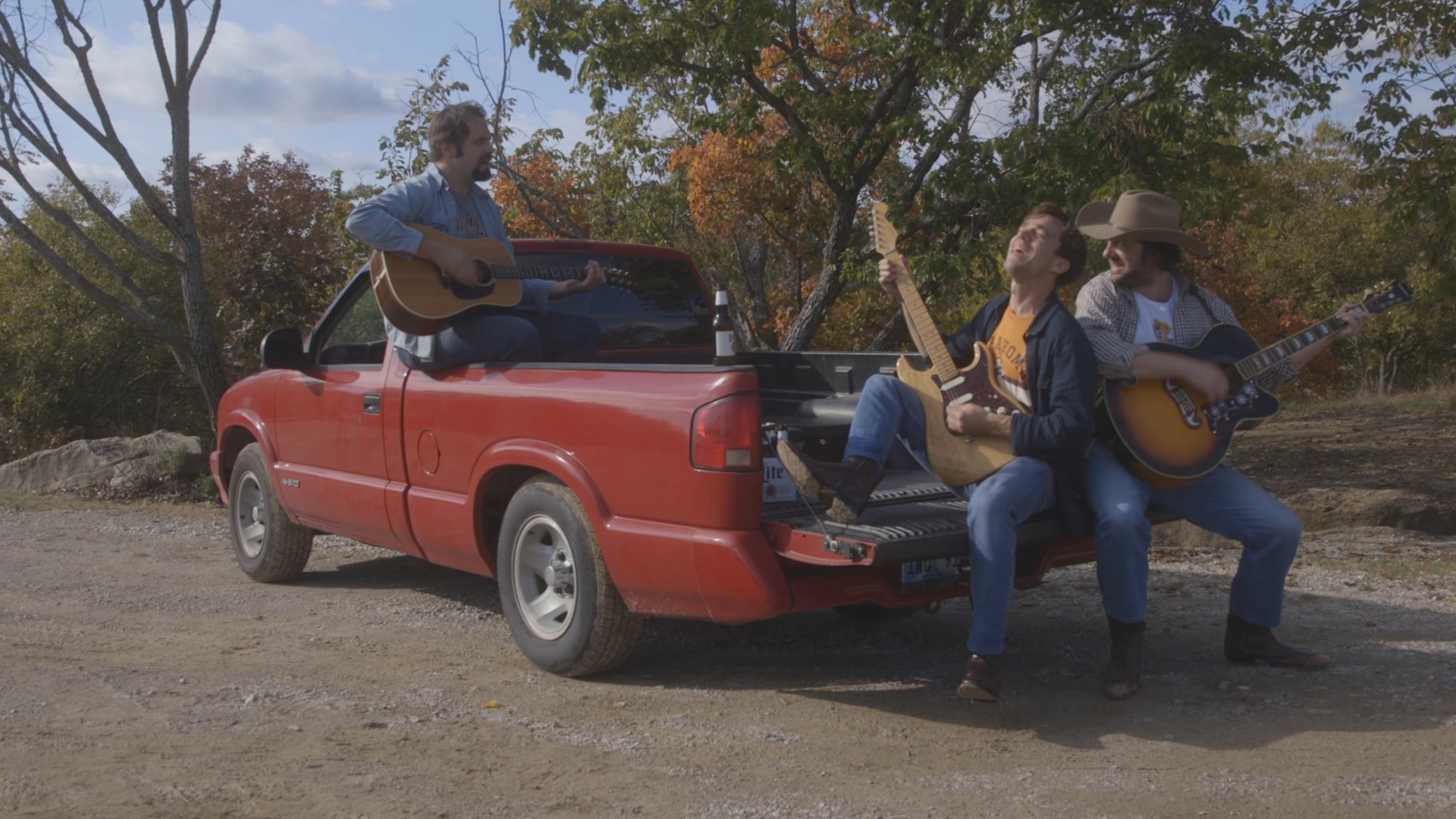 Three men, musicians, sit in the back of a red pickup truck parked on the side of a country lane