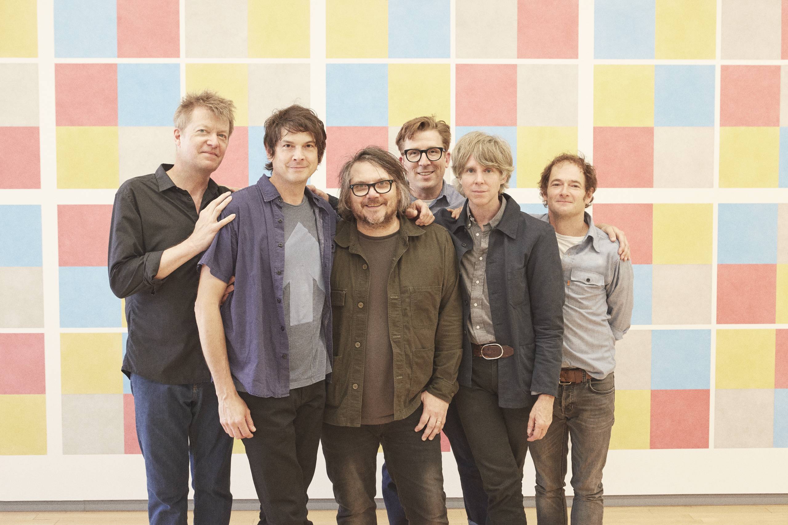 The members of the band Wilco standing in a row in front of a wall of colorful squares.