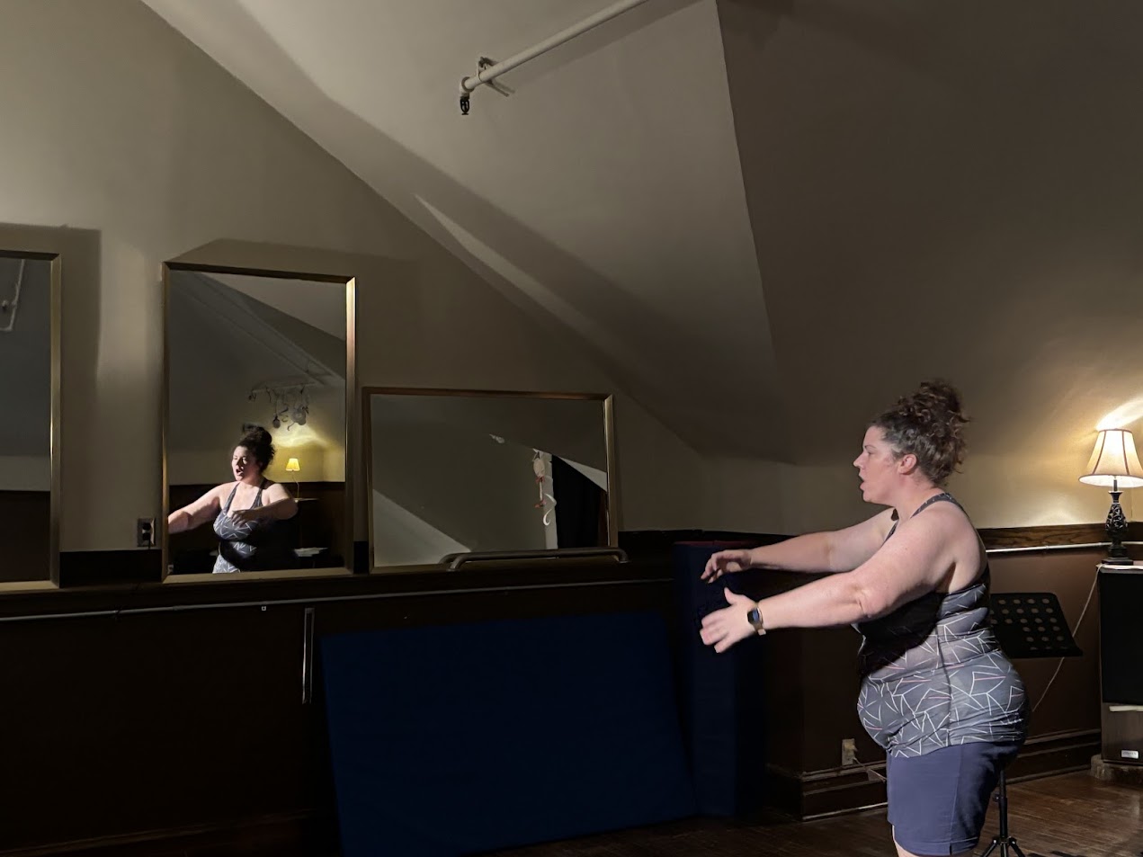 Person dancing in dim room with mirrored wall, music stand, and warm lamp lighting.