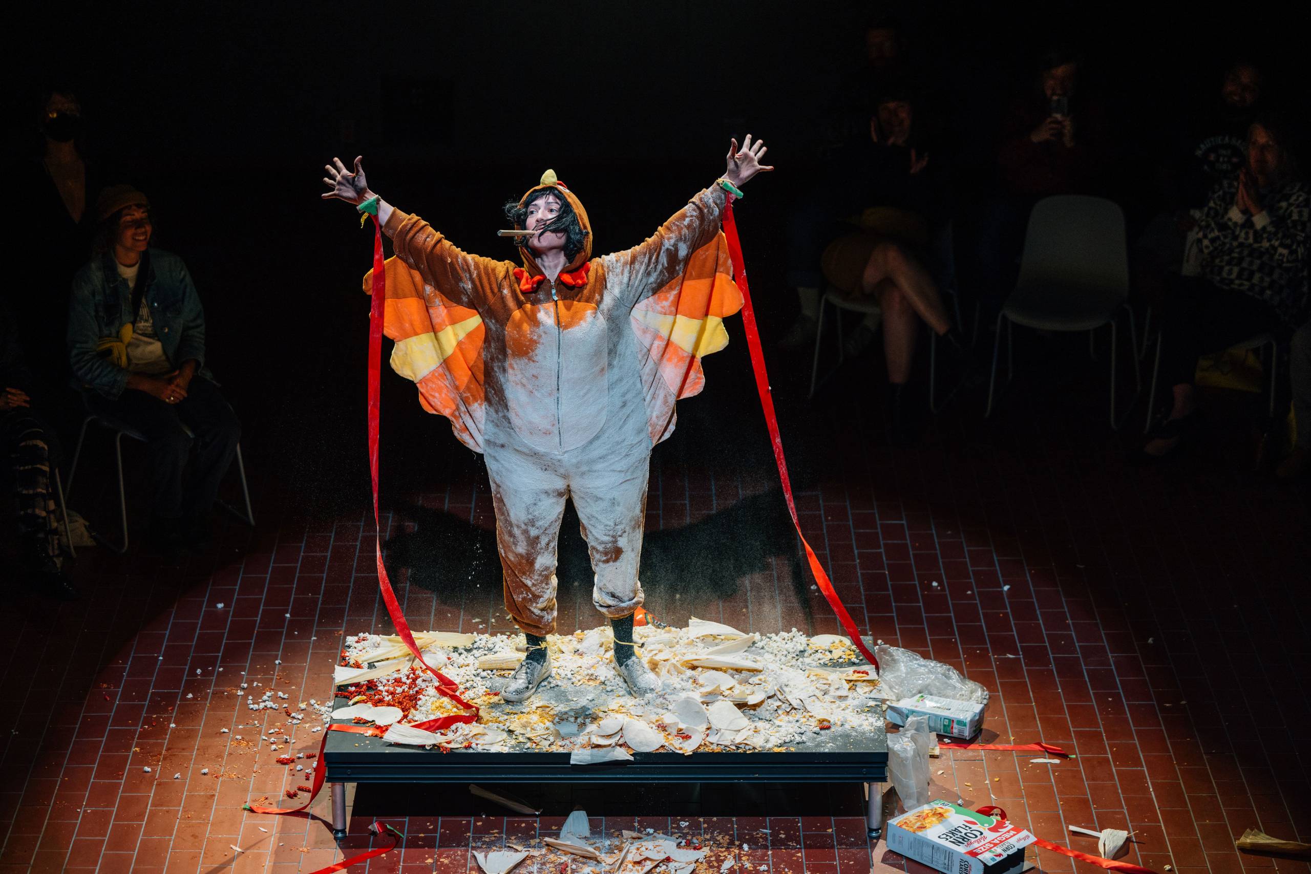 Artist Adriana Chavez stands on a small stage in a colorful chicken costume with her arms raised and a cigar in her mouth. Surrounding her are crushed corn chips, trash, and the remains of multiple Corn Flakes cereal boxes. Around her, just outside of the spotlight, sit audience members who are smiling and laughing.