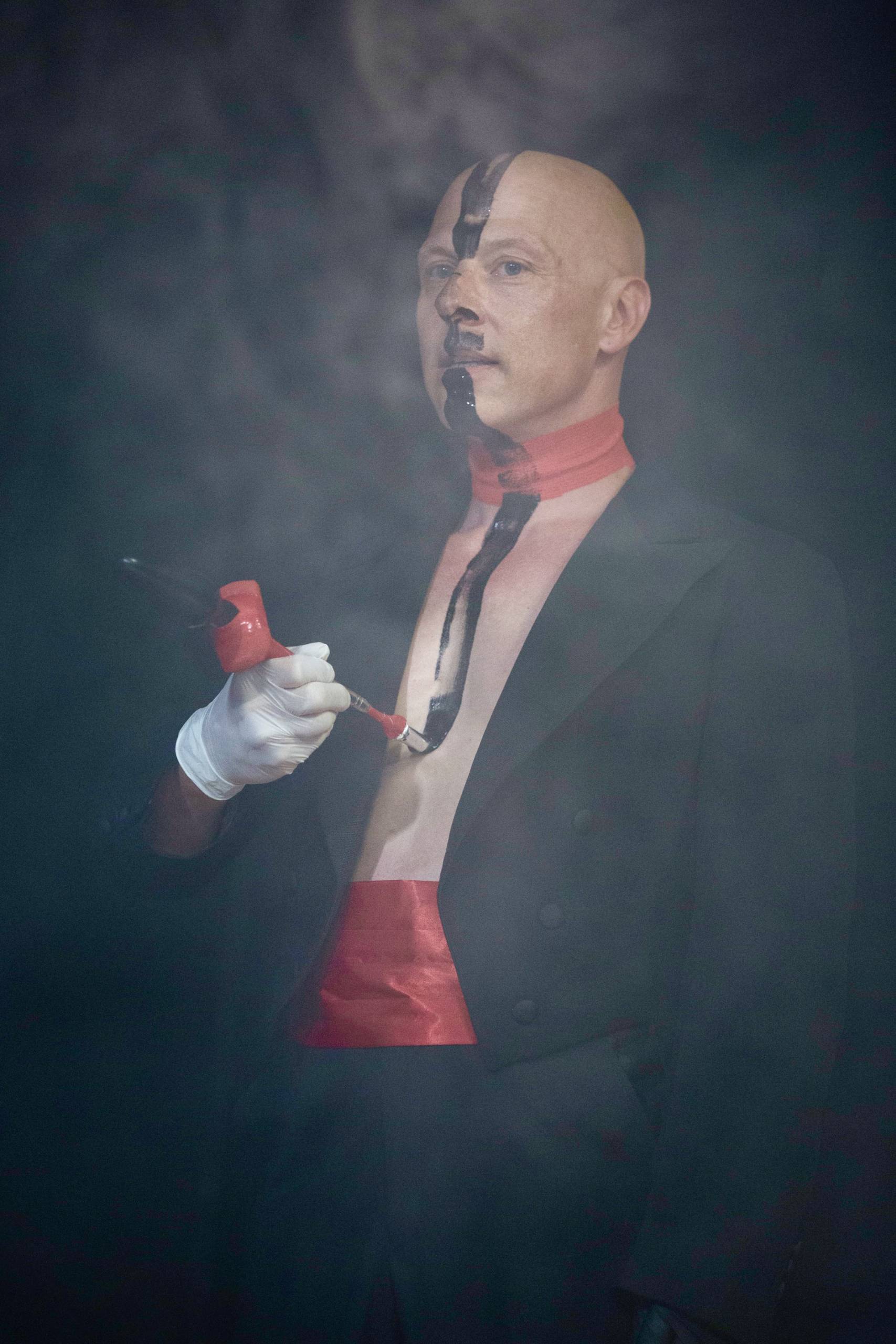 Bald person in formal attire painting face and chest black with a brush, red band on neck.