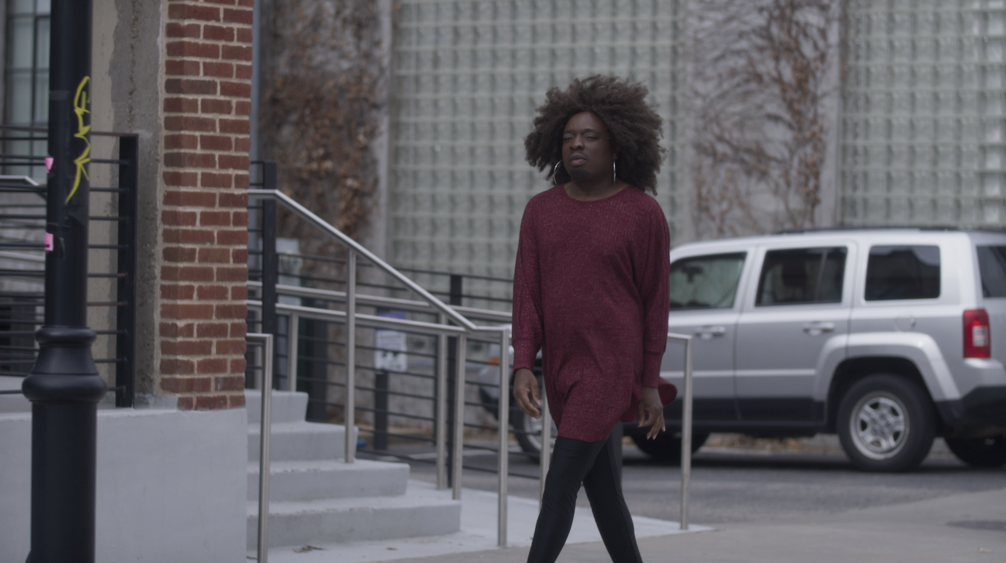 Person walking near a brick building and glass wall, silver SUV parked, black lamppost nearby.