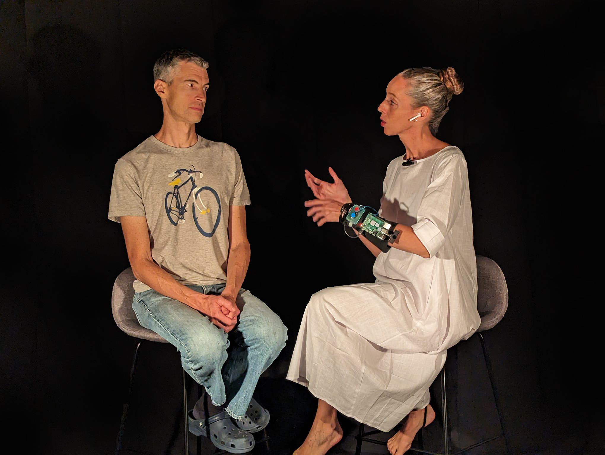 Man in bike t-shirt and woman in white with arm device seated on stools in dark room.