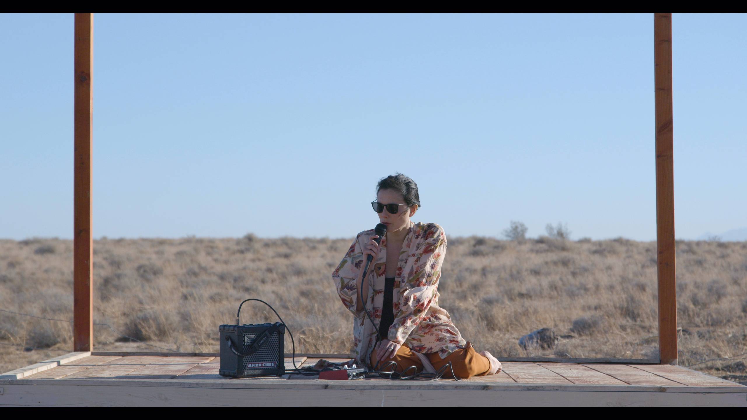 Person with microphone on wooden platform, dry open landscape, wearing sunglasses and floral outfit.