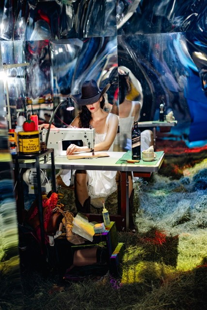 Person sewing in eclectic setting with a hat, reflective surfaces, and various colorful items.