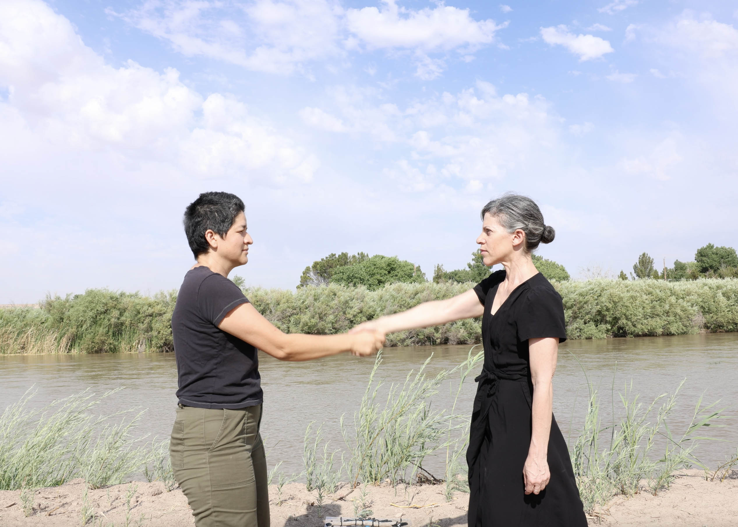 Two people shaking hands near a riverbank with bushes and trees under a partly cloudy sky.