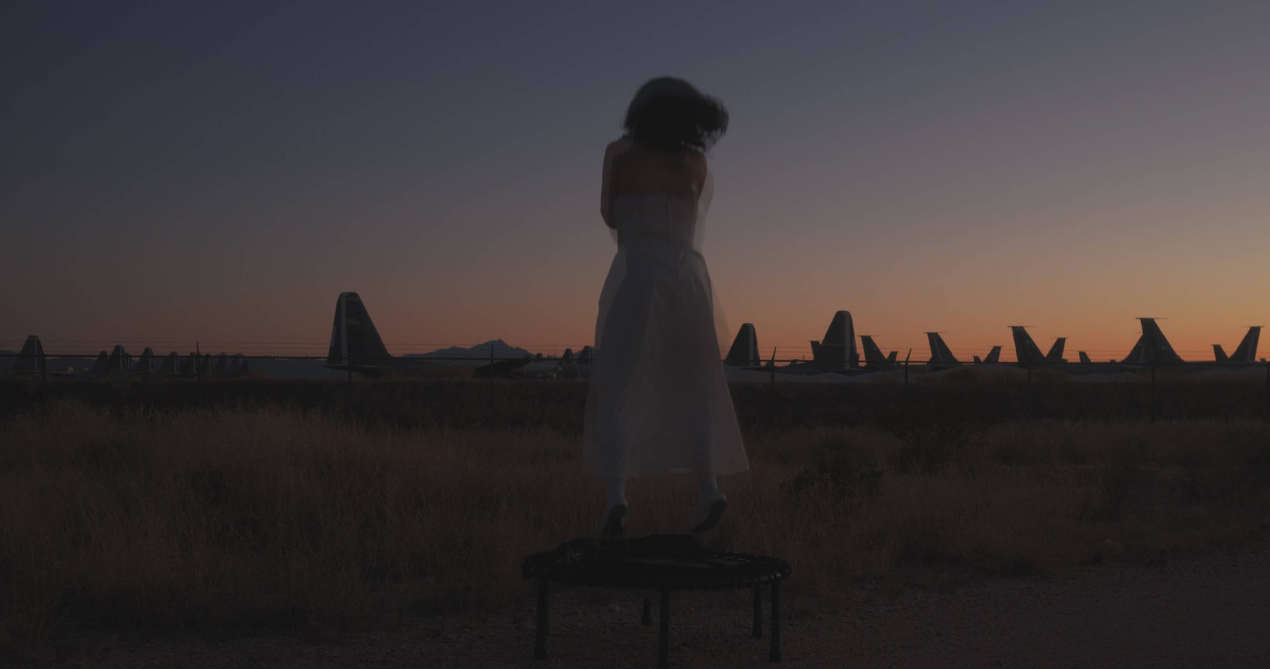 Person in white dress on trampoline, aircraft tails silhouetted at dusk in the background.