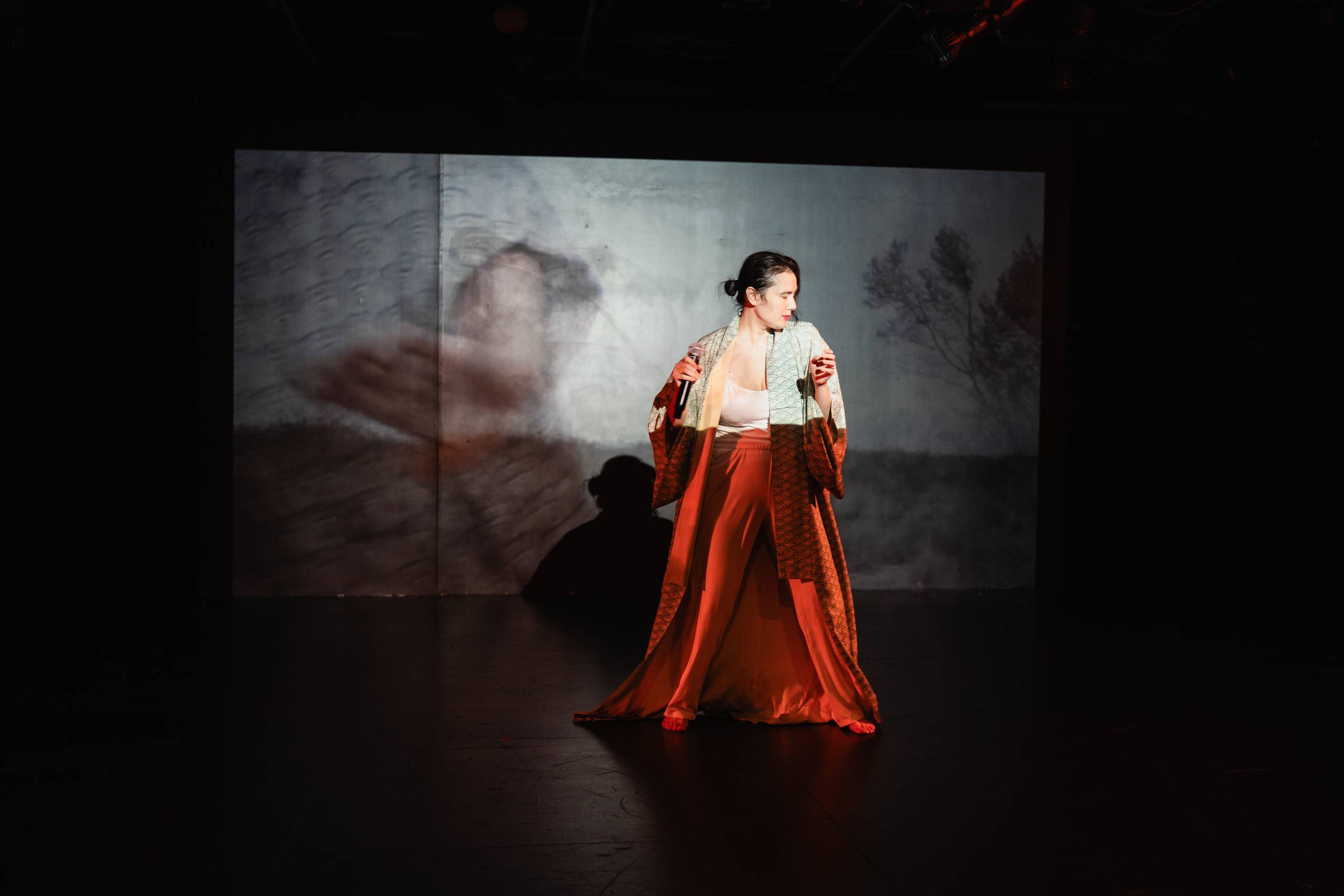 Person on stage in robe and orange skirt, abstract art backdrop with tree silhouettes.