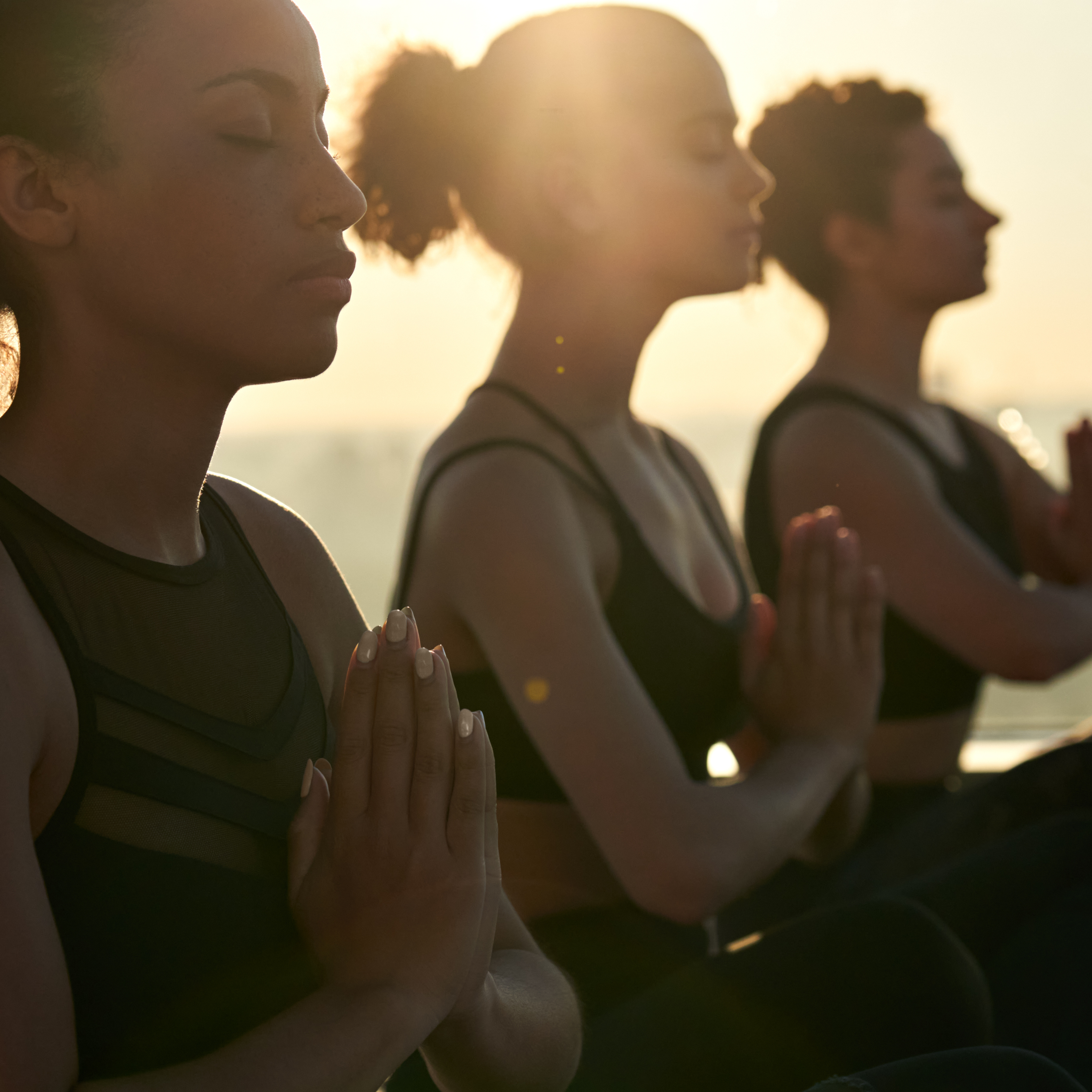 Three women in black workout outfits sit crosslegged, their hands pressed together in a namaste position