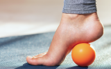 A woman's foot raised onto its toes, with an orange ball under the arch of the foot.