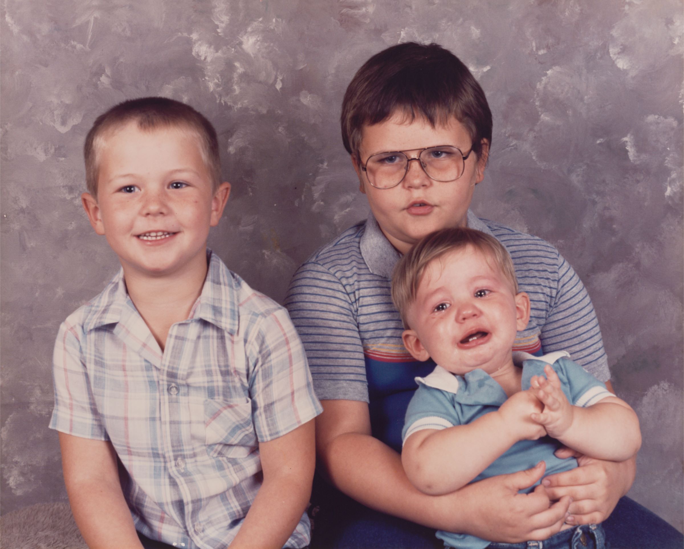 A family portrait showing three young boys, two between the ages of 7 and 10, and one who is a toddler. The boys sit in front of a marbled grey portrait studio background.
