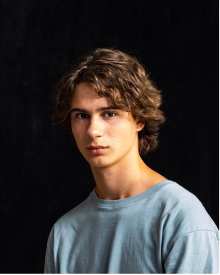 Unno Young person with curly hair in light blue shirt against black background, looking at camera.