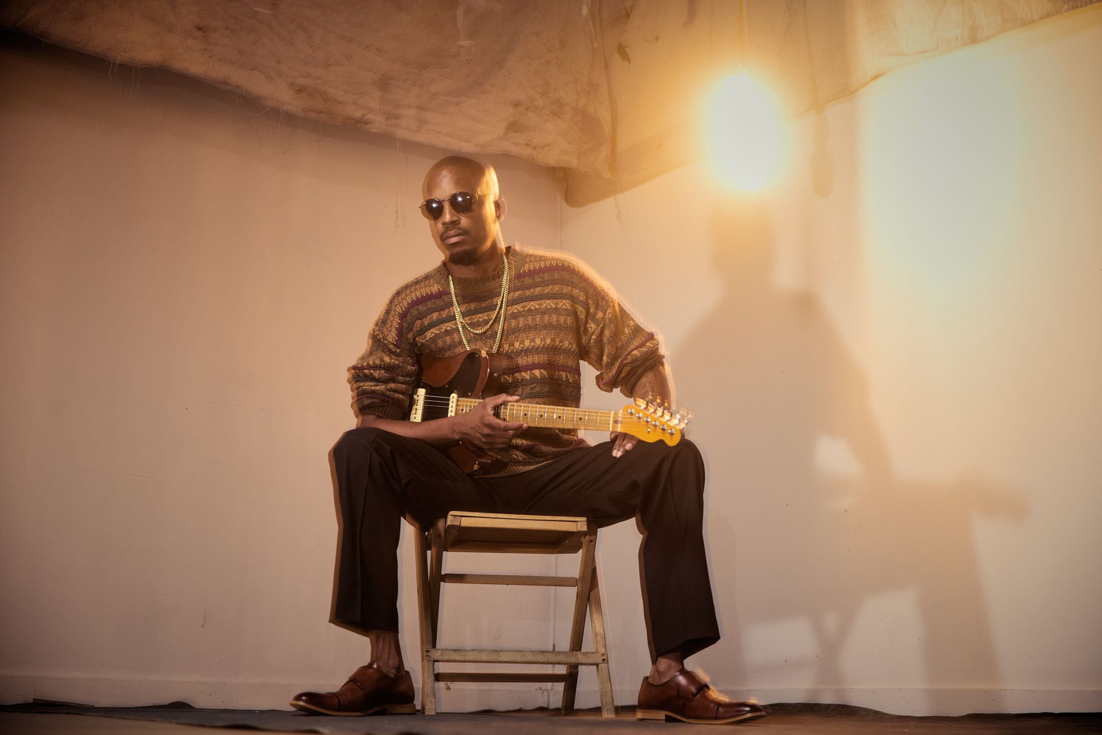 Soul and Blues musician Black Joe Lewis sitting on a chair with a guitar in front of an empty corner