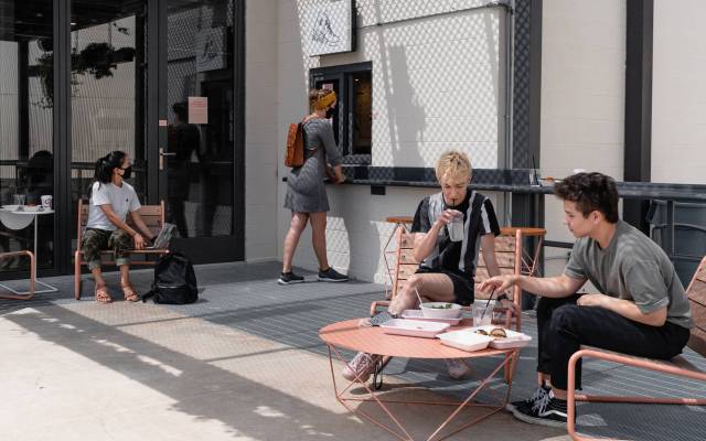 Zona de patio con grupos de personas con bebidas, una mujer al fondo está en una ventanilla de servicio